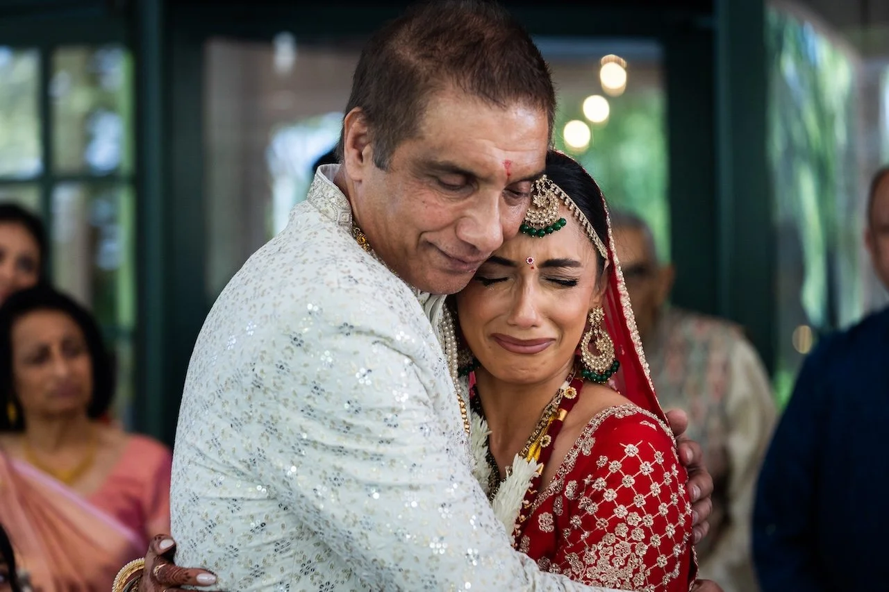 A man and woman are hugging during what appears to be a wedding, with emotional expressions. The woman is dressed in traditional Indian wedding attire with jewelry, and the man is wearing a white, embroidered outfit. Other guests are visible in the b