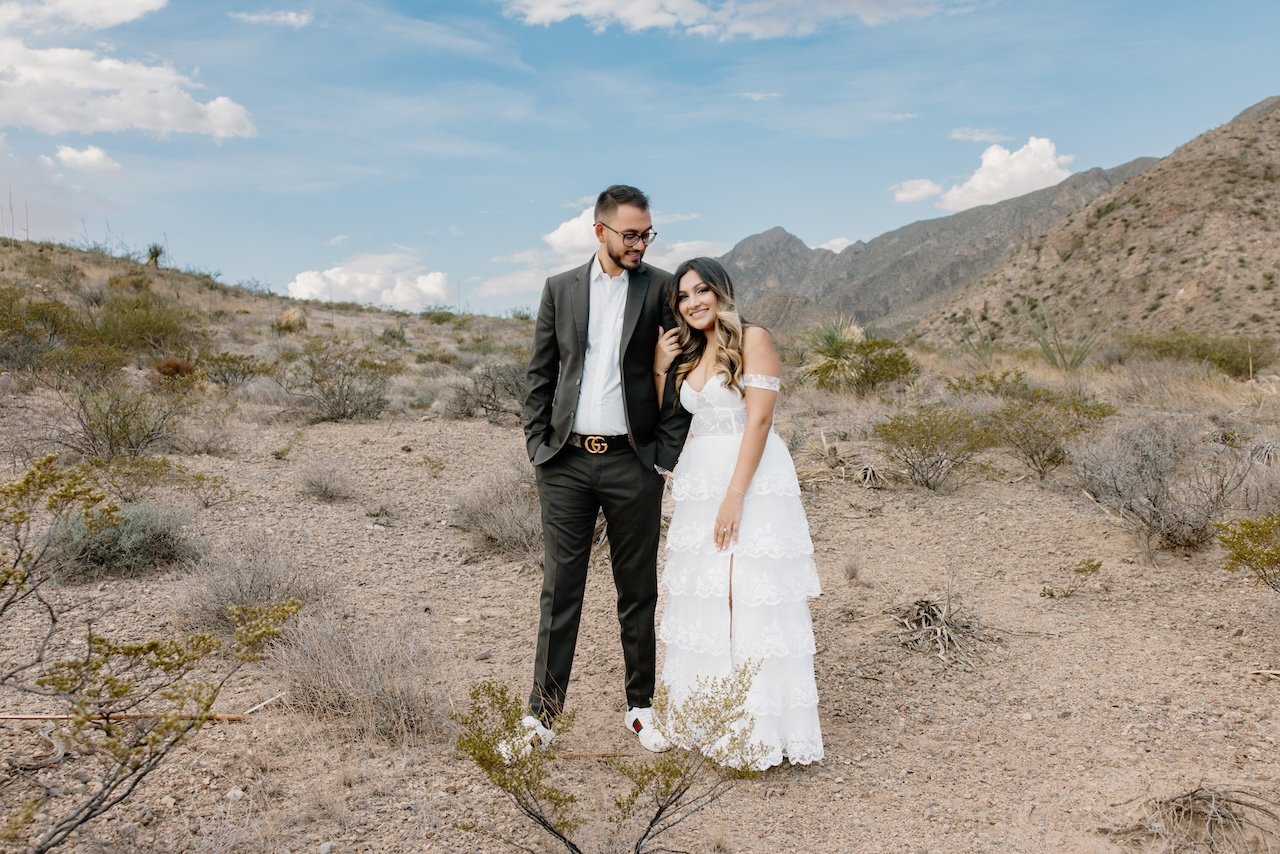 A couple dressed in wedding attire standing in a desert with mountains in the background, the man in a black suit and the woman in a white lace dress.