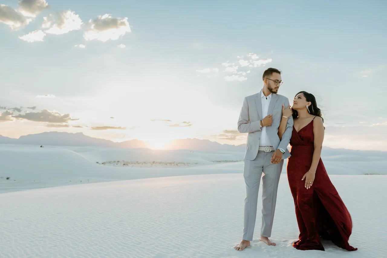 A man and woman in formal attire standing in a desert landscape with sand dunes during sunset.