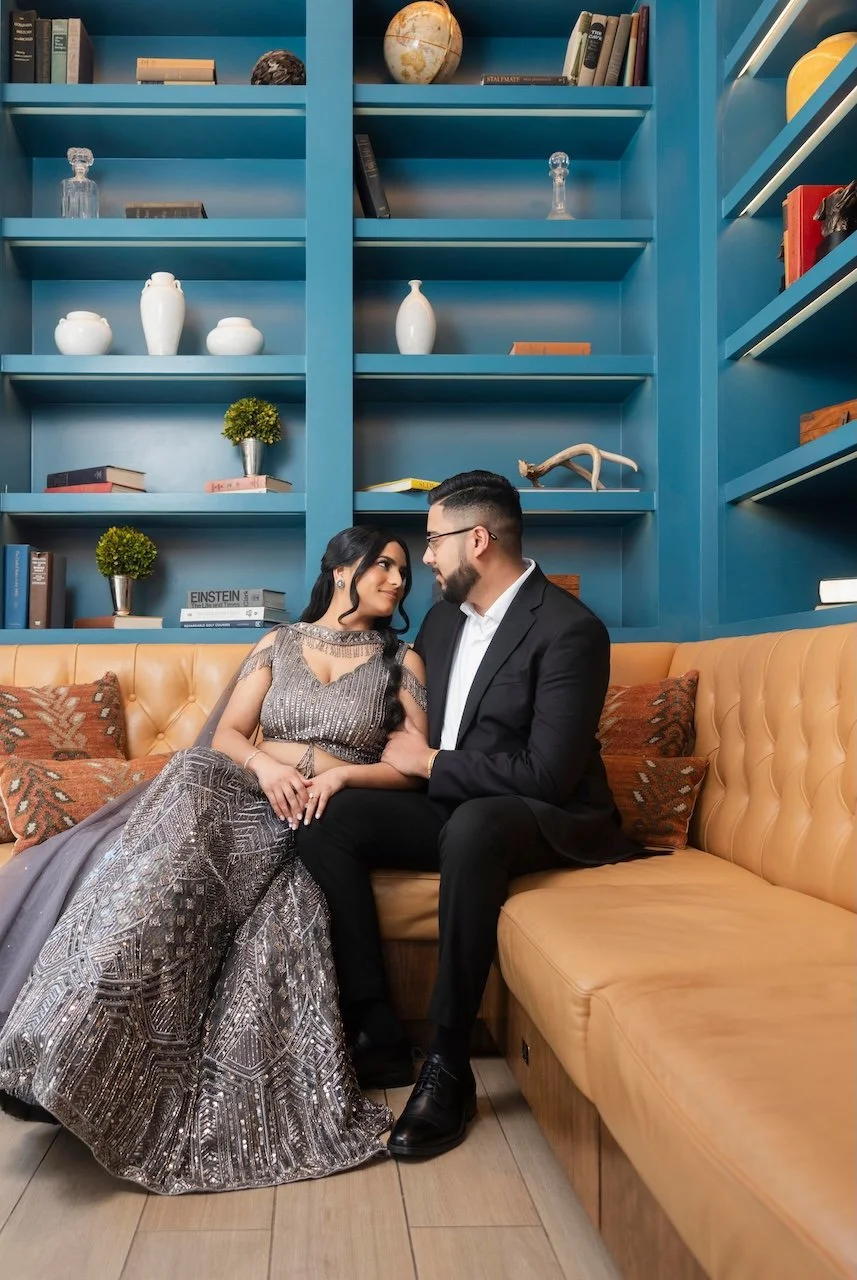 A couple sitting on a mustard-colored sofa, gazing into each other's eyes, with a blue bookshelf in the background decorated with books, vases, a globe, and sculptures.