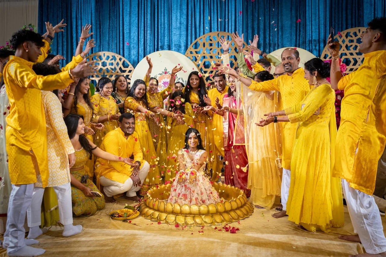 A group of people dressed in yellow and traditional Indian attire celebrating around a woman sitting in a decorated small pond, with flower petals and festive decorations in the background.
