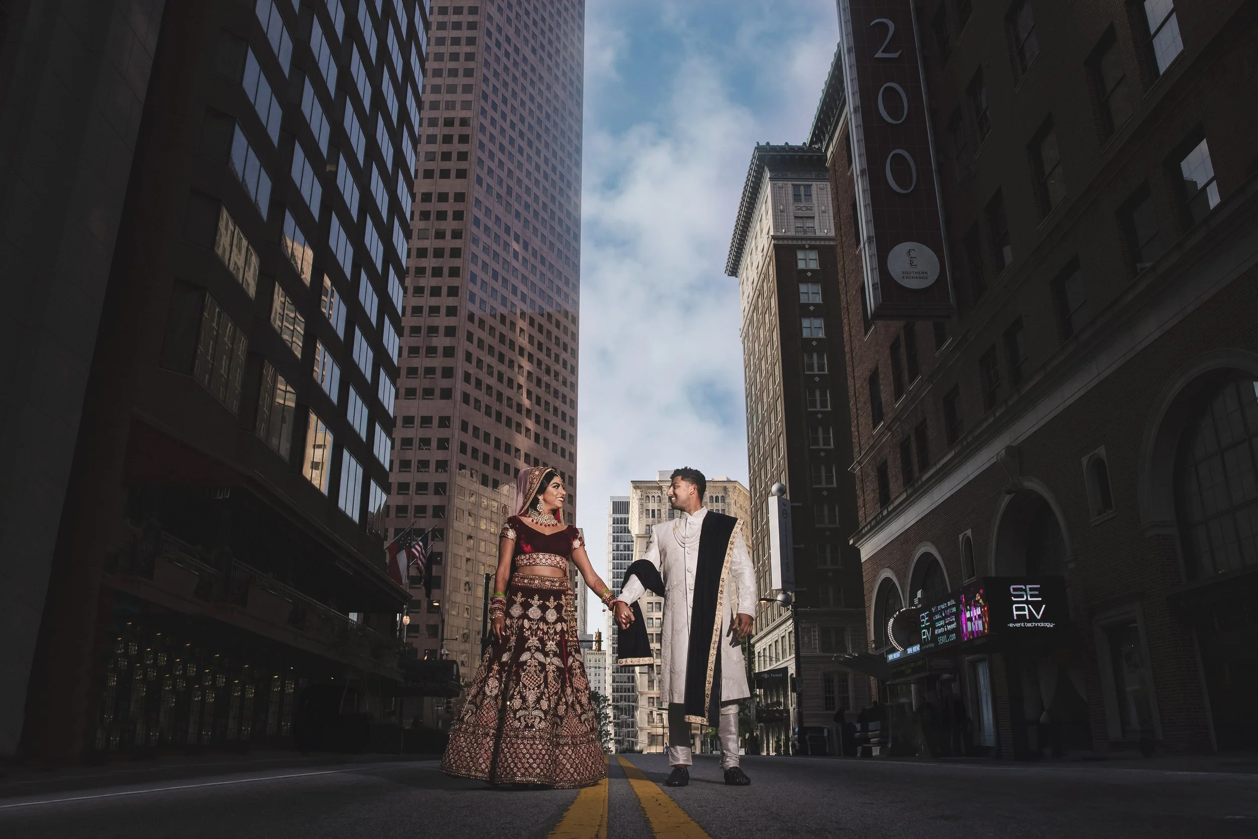 A couple in traditional Indian wedding attire standing in the middle of a city street, holding hands and looking at each other, surrounded by tall skyscrapers.