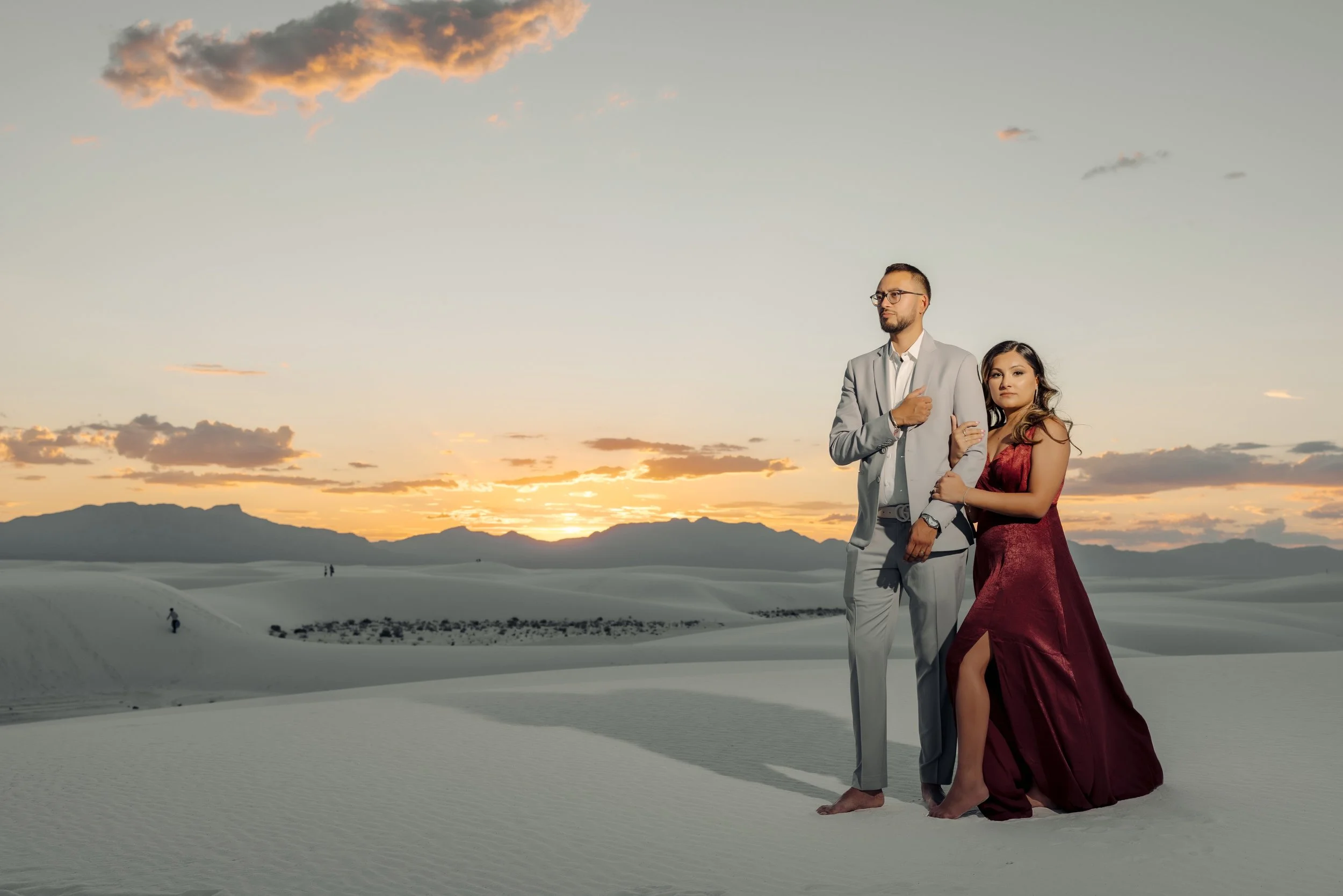 A man and woman standing close together in a desert landscape at sunset, with mountains in the background. The man is wearing a light-colored suit and glasses, while the woman is wearing a long, burgundy dress with a slit. The woman is holding the ma