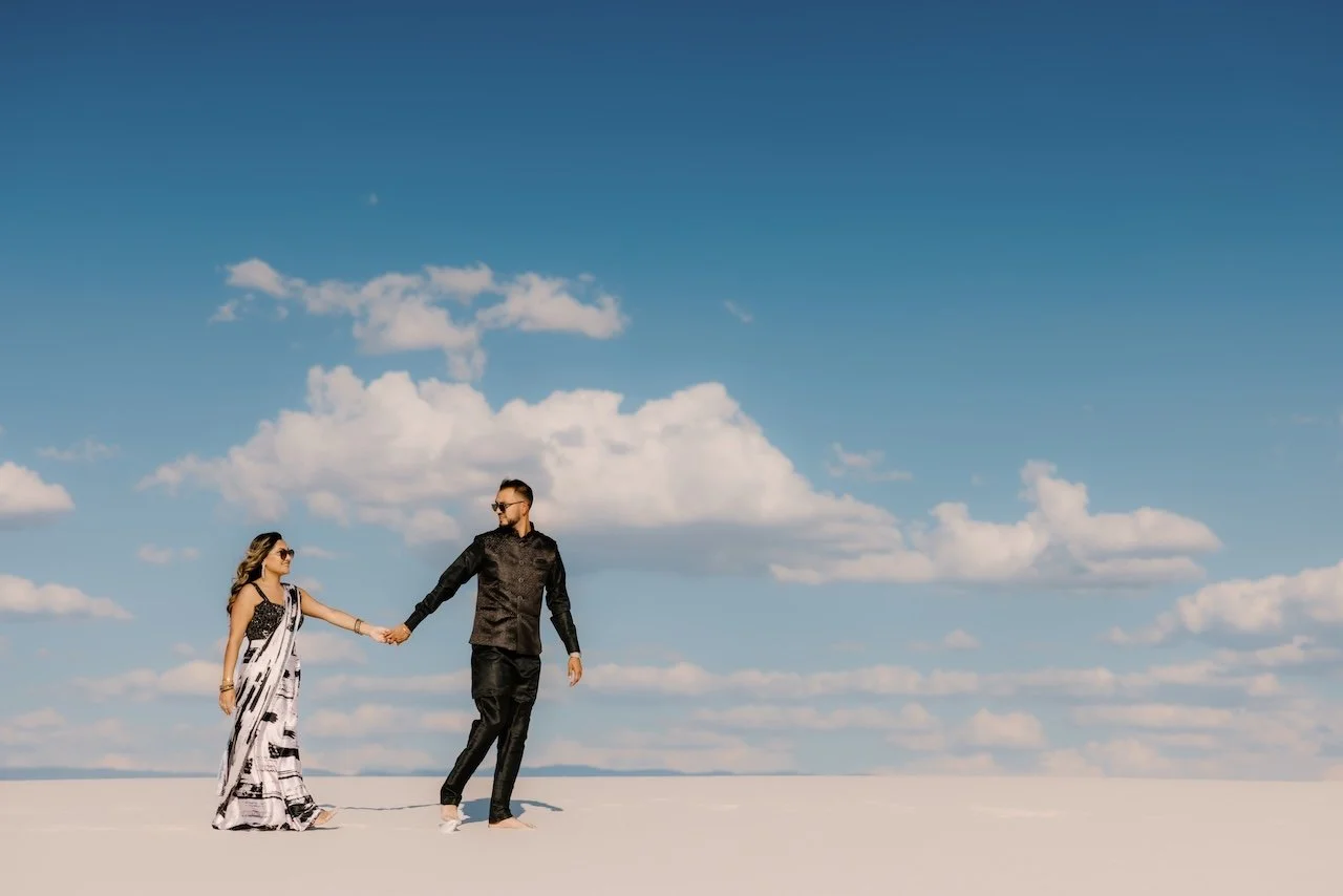 A couple holding hands and walking on a white desert landscape under a blue sky with clouds.