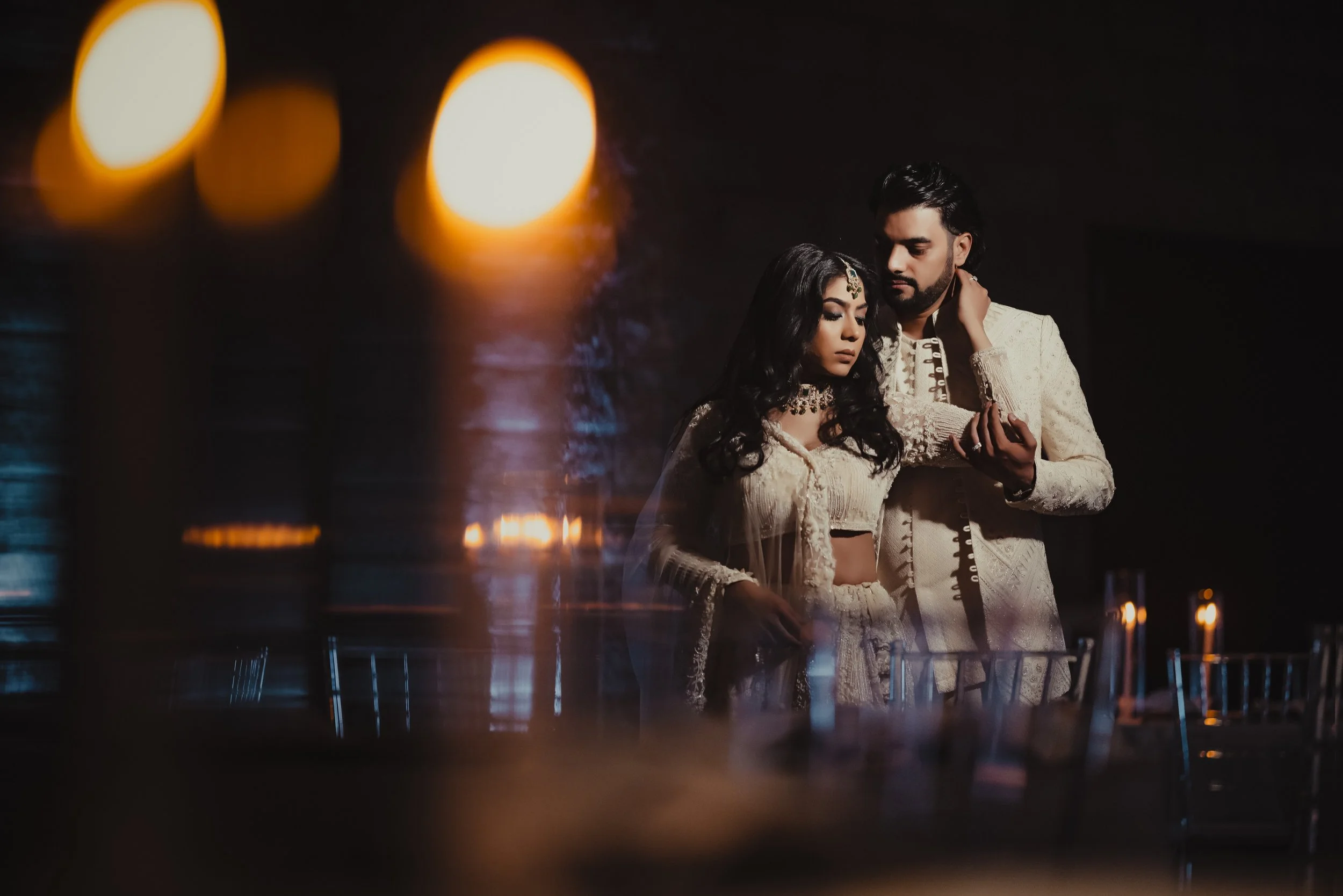 A couple dressed in traditional Indian attire standing close together in a dimly lit setting, with warm candlelight reflections and blurred lights in the background.