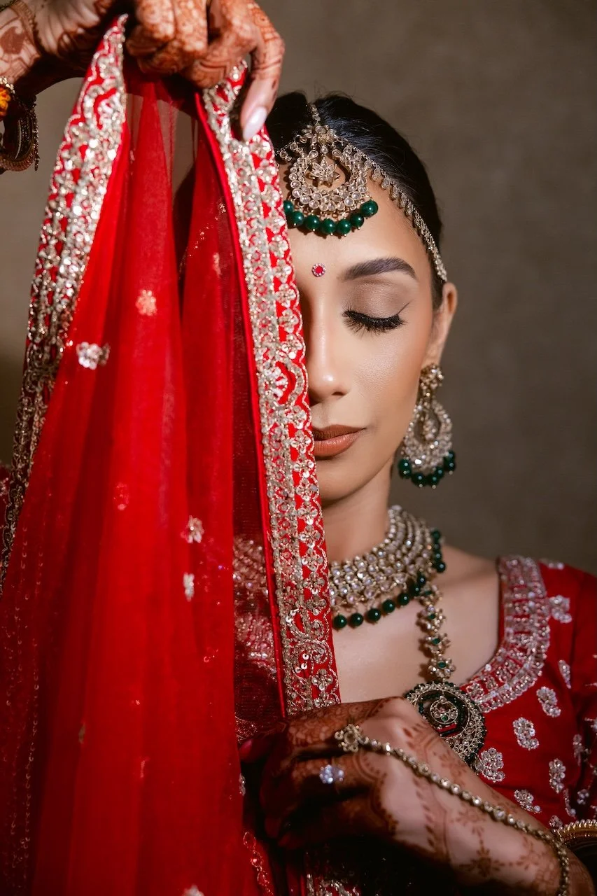Young woman dressed in traditional red Indian bridal attire with detailed jewelry, henna on her hand, holding the edge of her dupatta, with her eyes closed.