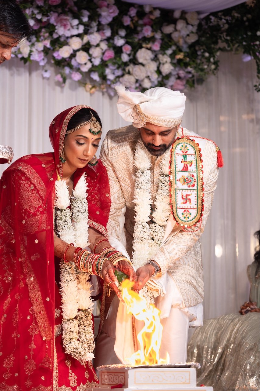Indian couple in traditional wedding attire performing a fire ritual during their wedding ceremony, with floral decorations in the background.