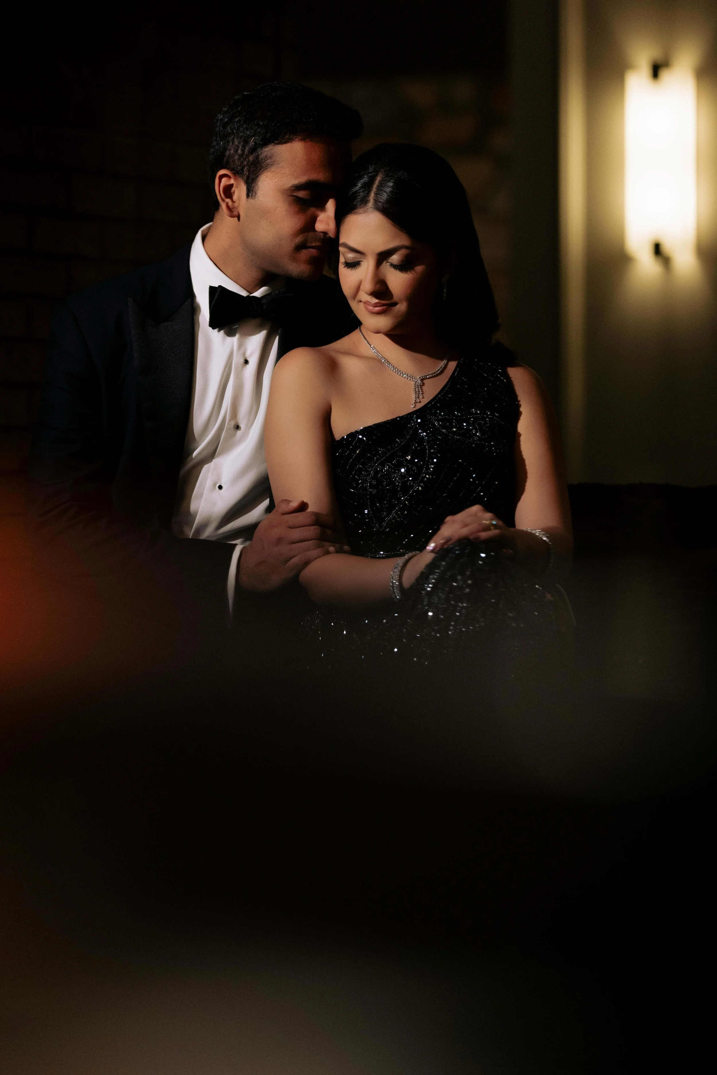 A man in a tuxedo and a woman in a black evening gown sit close together, with the man gently holding the woman's arm, in a dimly lit setting.