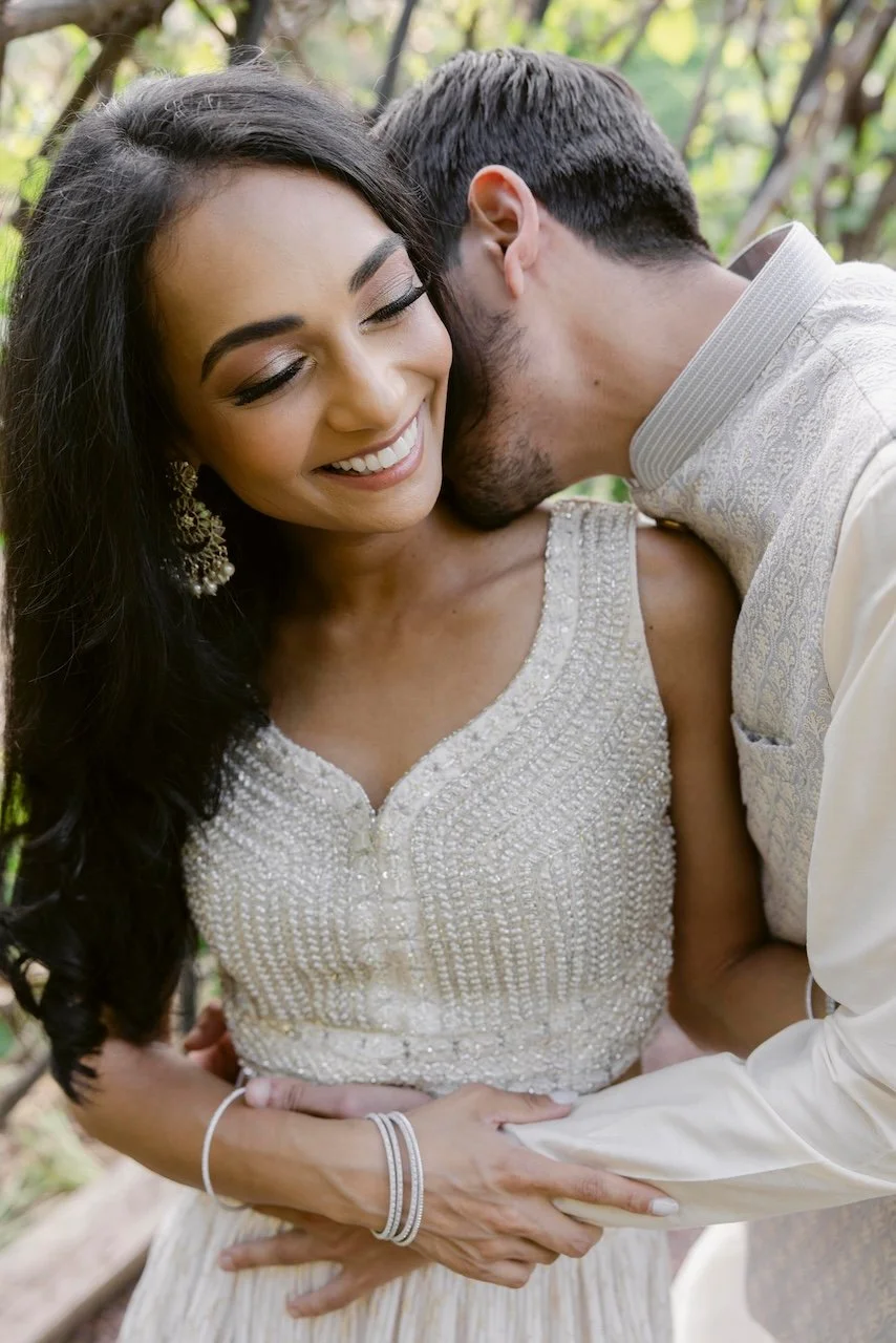 A couple embracing outdoors, woman smiling with eyes closed, man kissing her cheek, trees in background, woman wearing traditional Indian attire with jewelry.
