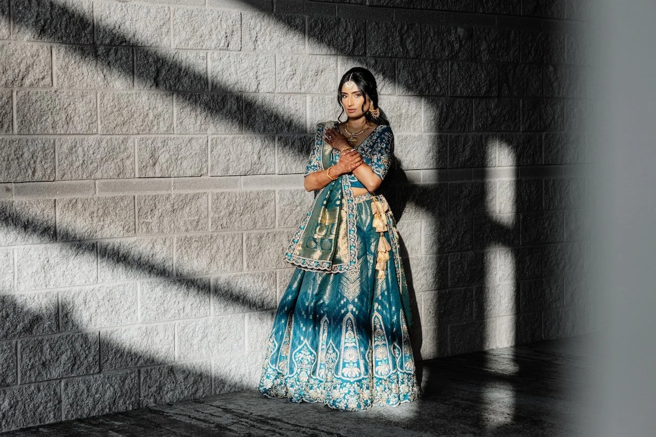 Woman wearing traditional Indian attire with jewelry, standing against a brick wall with shadow patterns.