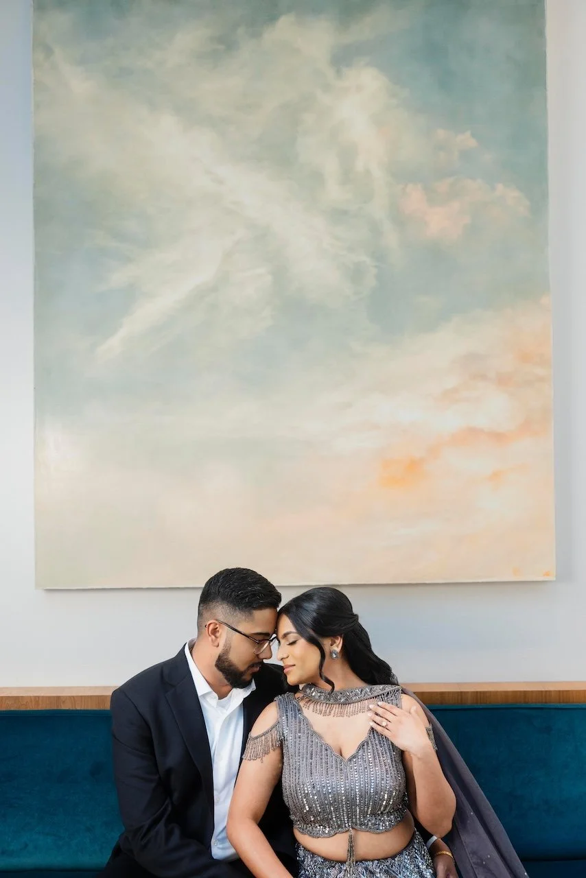 A man and woman sitting closely with foreheads touching, dressed in formal attire, in front of a large painting of a cloudy sky.
