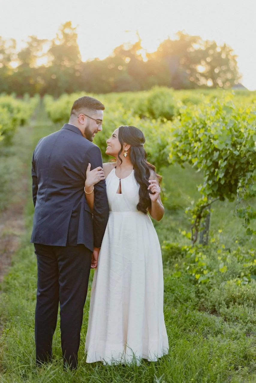 A couple dressed in wedding attire standing in a lush vineyard at sunset, gazing into each other's eyes and smiling.