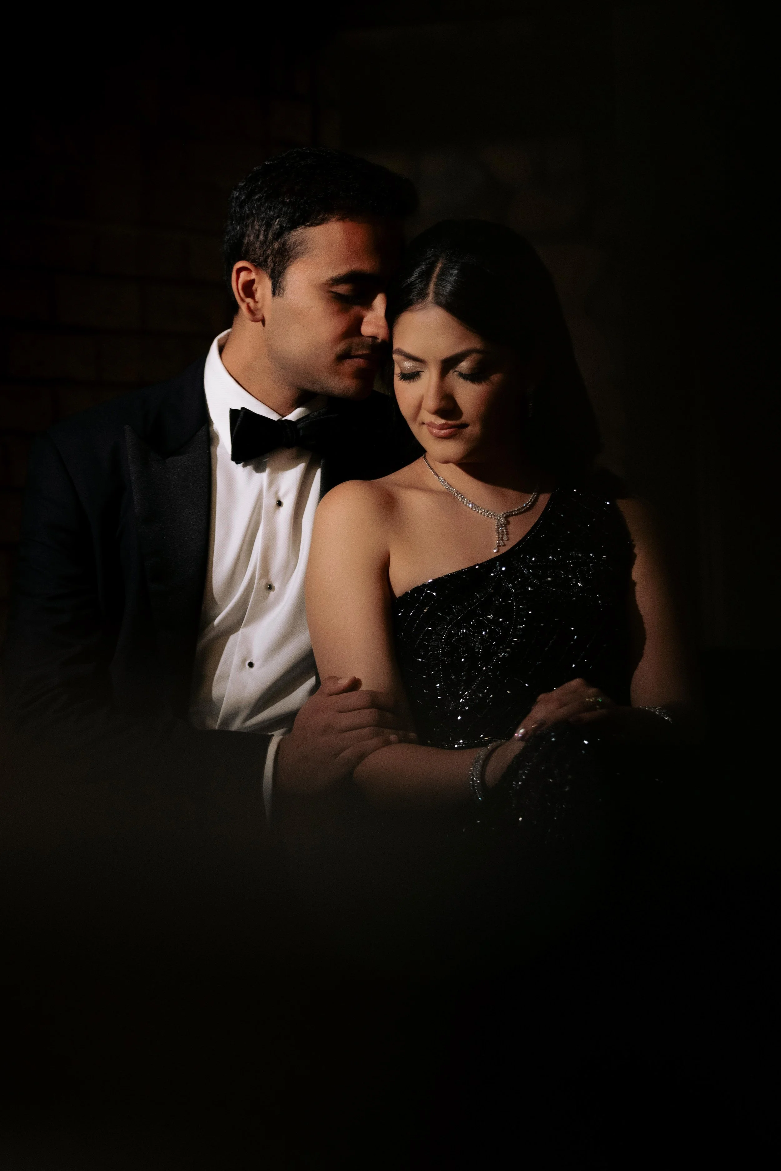 A man and woman in formal attire embrace closely in a dark setting, with the man's face touching the woman's forehead, both with eyes closed.