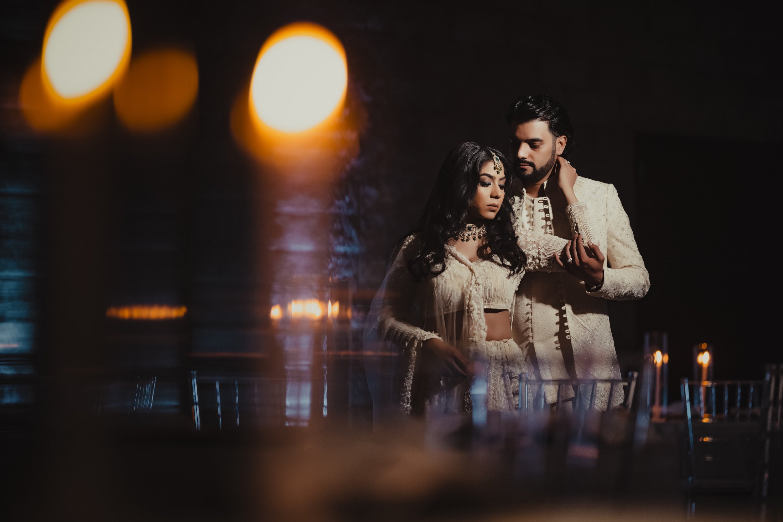 A man and woman dressed in traditional Indian attire, standing close together in a dark, ambient setting with candlelight reflections and blurred candles in the foreground.