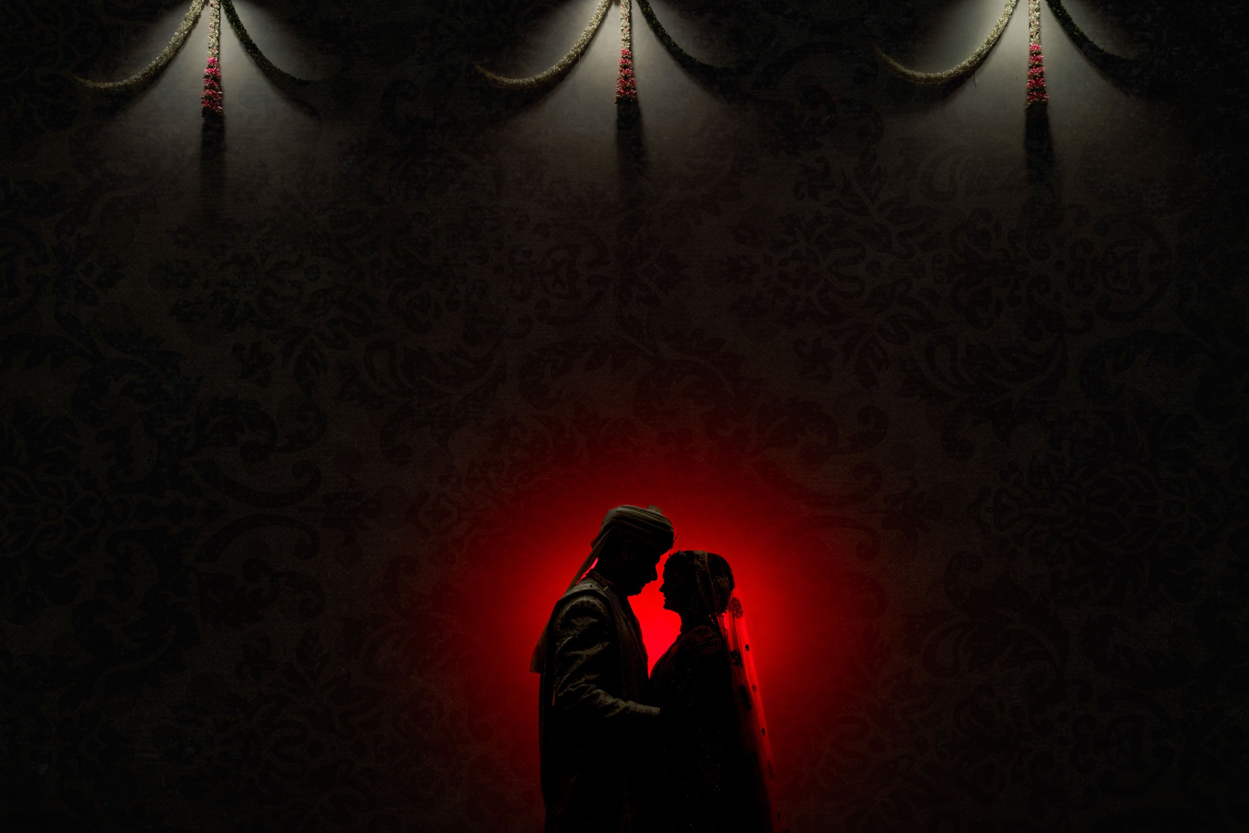 Silhouette of a couple, possibly a bride and groom, touching foreheads with a red glow behind them, against a dark background with ornate patterns and a decorative curtain at the top.