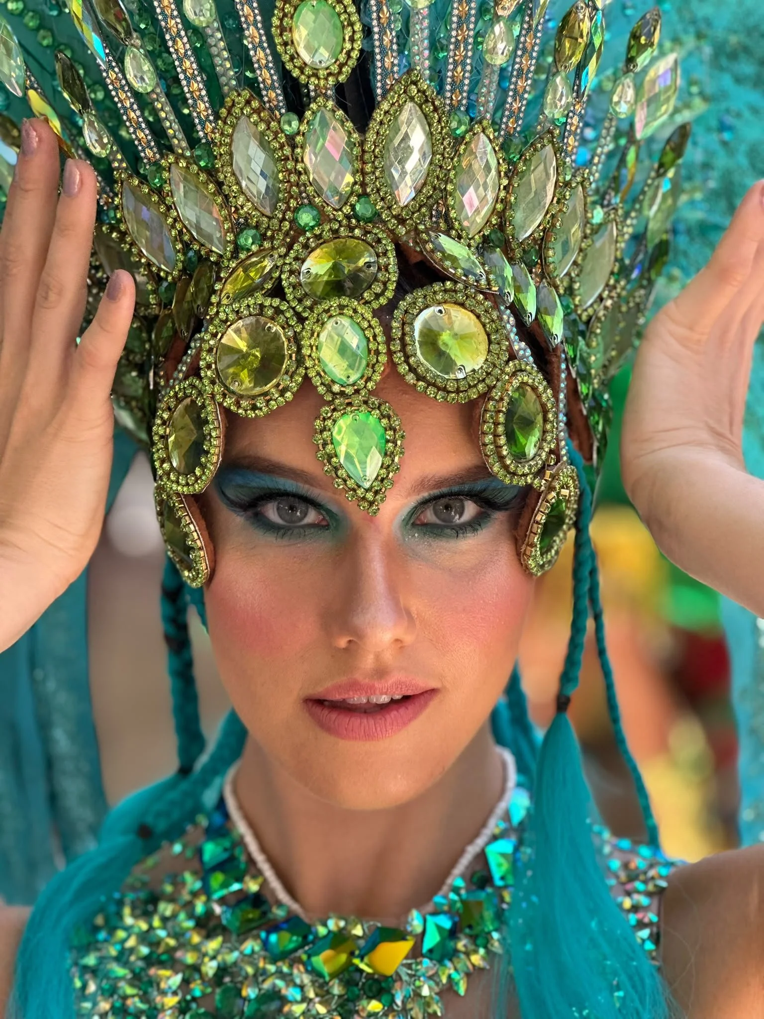 A woman wearing a colorful, jewel-encrusted costume with a large, ornate headdress adorned with green and clear gemstones. She has light makeup with green eye shadow and is holding the headdress with both hands.