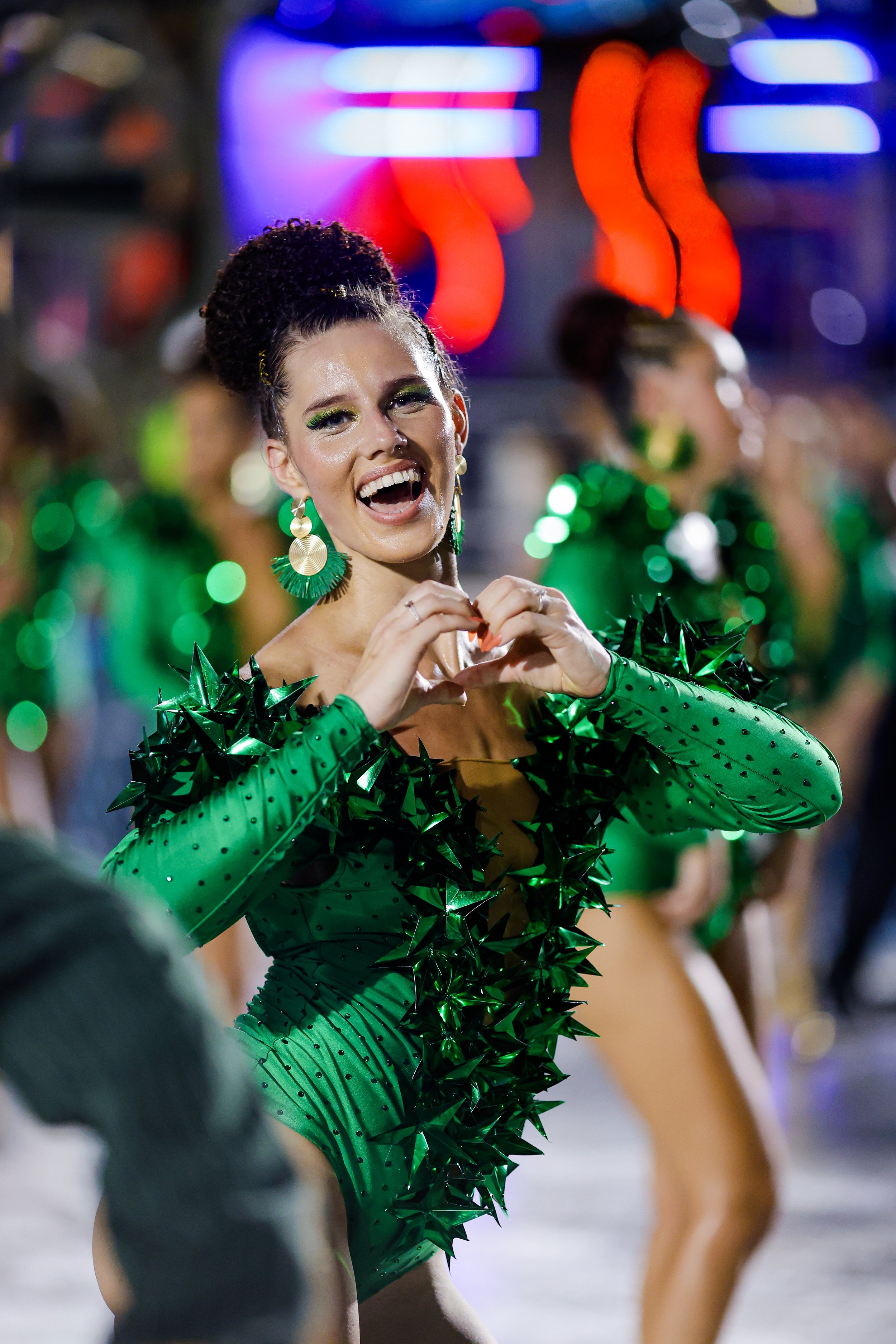 A woman in a green costume with shiny star embellishments and large circular earrings is smiling and making a heart shape with her hands. She is participating in a parade or performance, with other similarly dressed women behind her and colorful lights in the background.