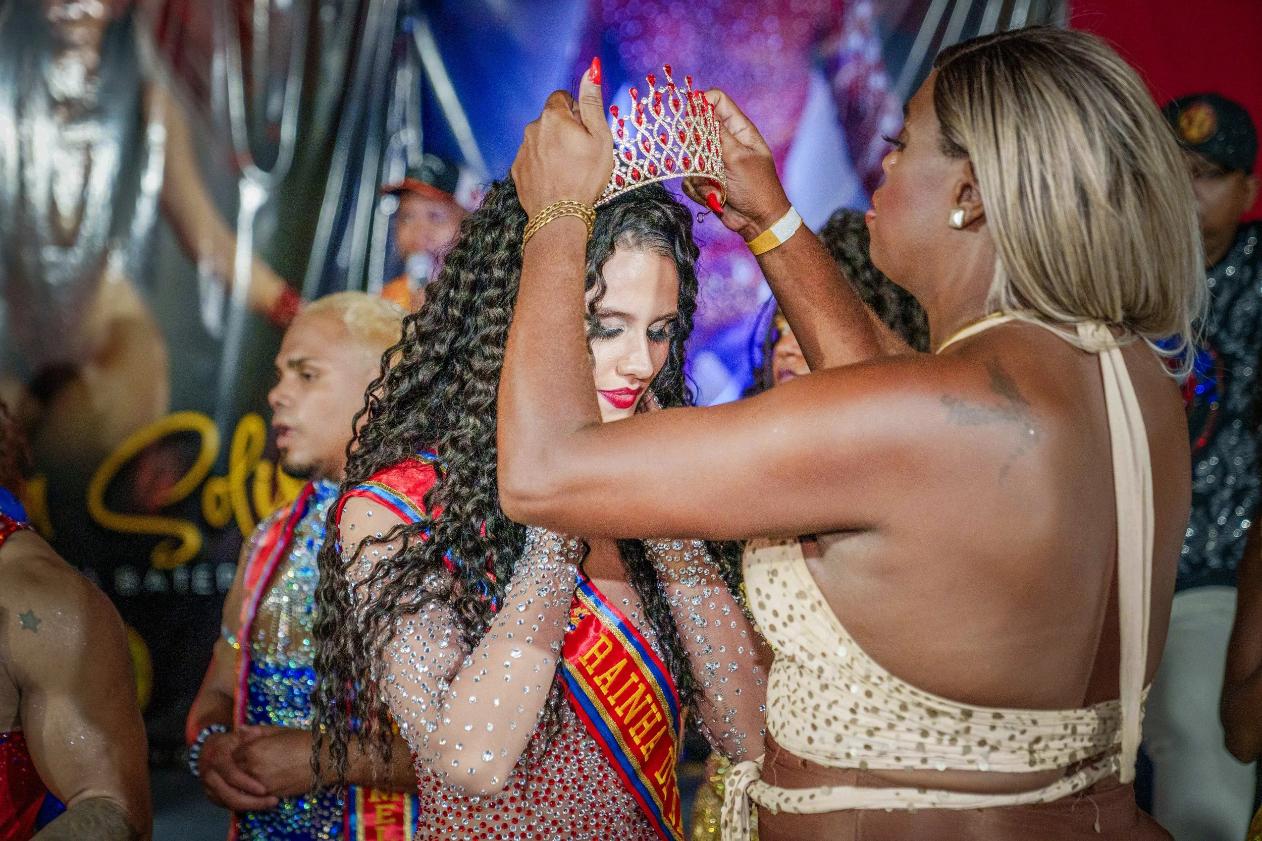 A woman with long curly dark hair is being crowned with a jeweled tiara by another woman. The woman receiving the crown has a sash across her chest, and both women are dressed in shiny, decorated clothing, likely participating in a celebration or pageant.