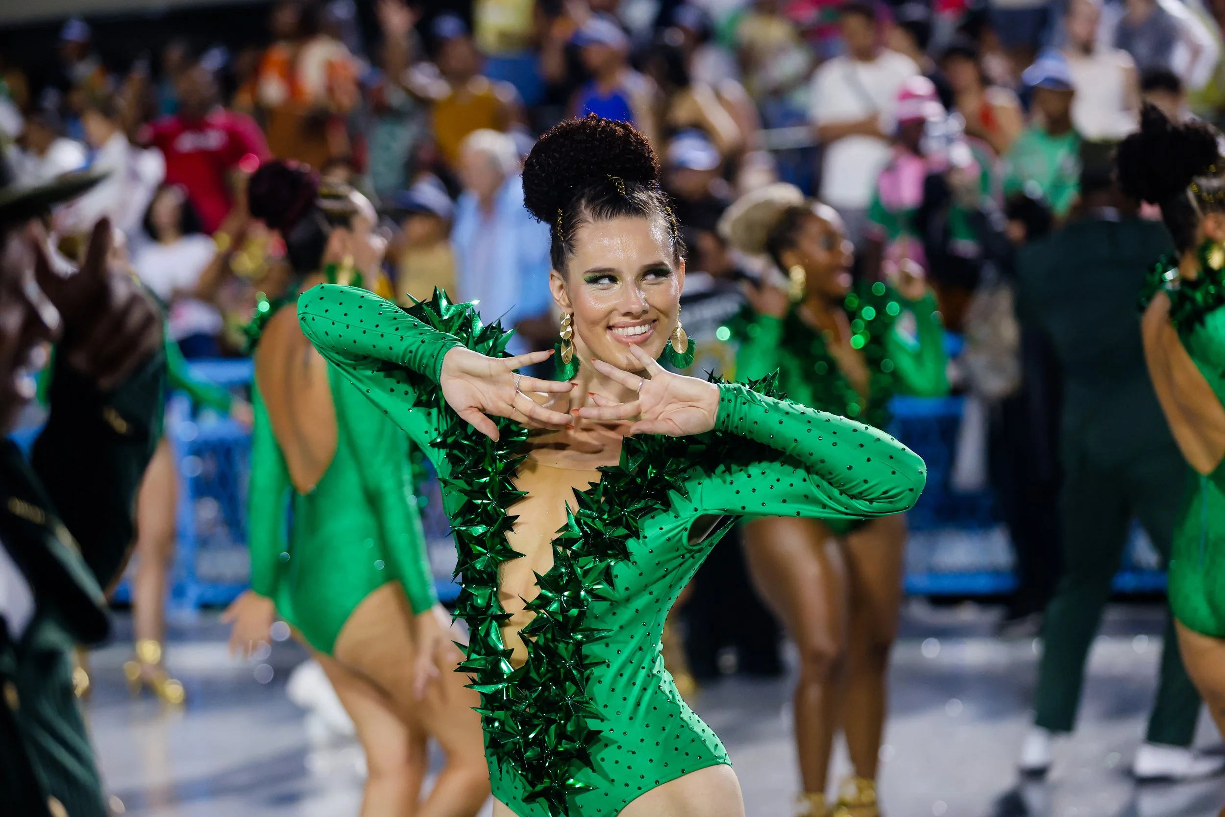 A woman dressed in a vibrant green costume with black polka dots and spiked decorative elements performs a dance during a parade, with a large crowd of onlookers in the background.