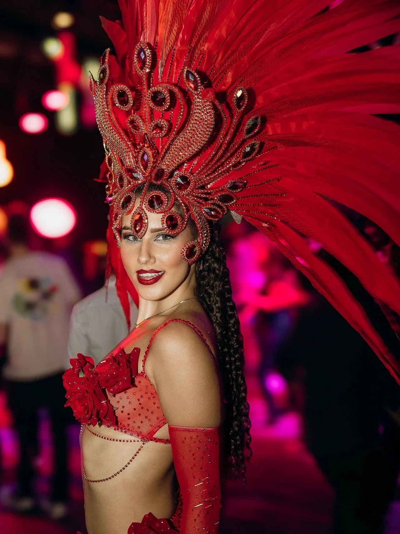 A woman wearing an elaborate red costume with feathered headdress and rhinestones, smiling at the camera, at a festive event.