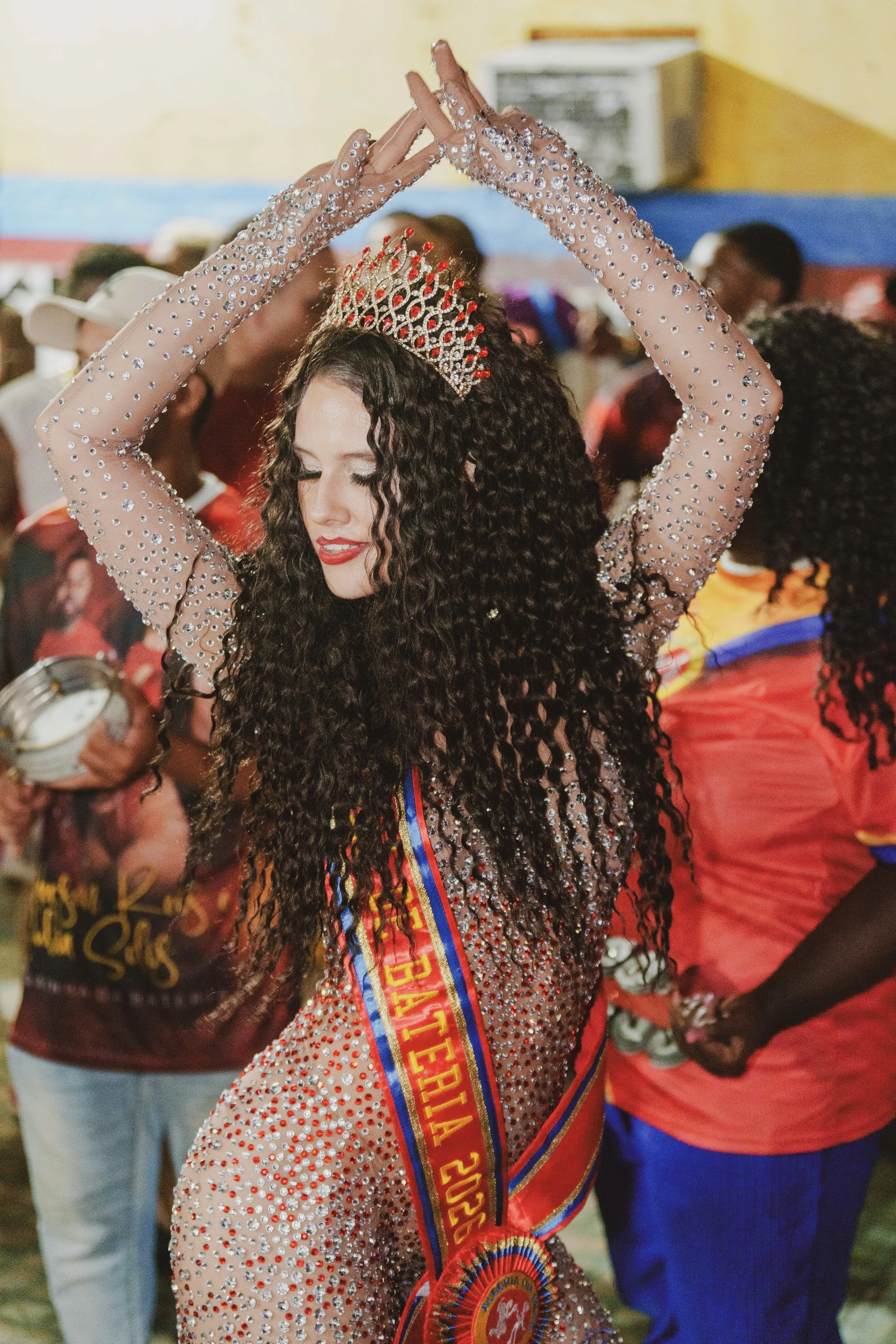A woman with long curly hair wearing a crown and a dress covered in rhinestones, dancing with her arms raised above her head. She is wearing a sash and a large medal.