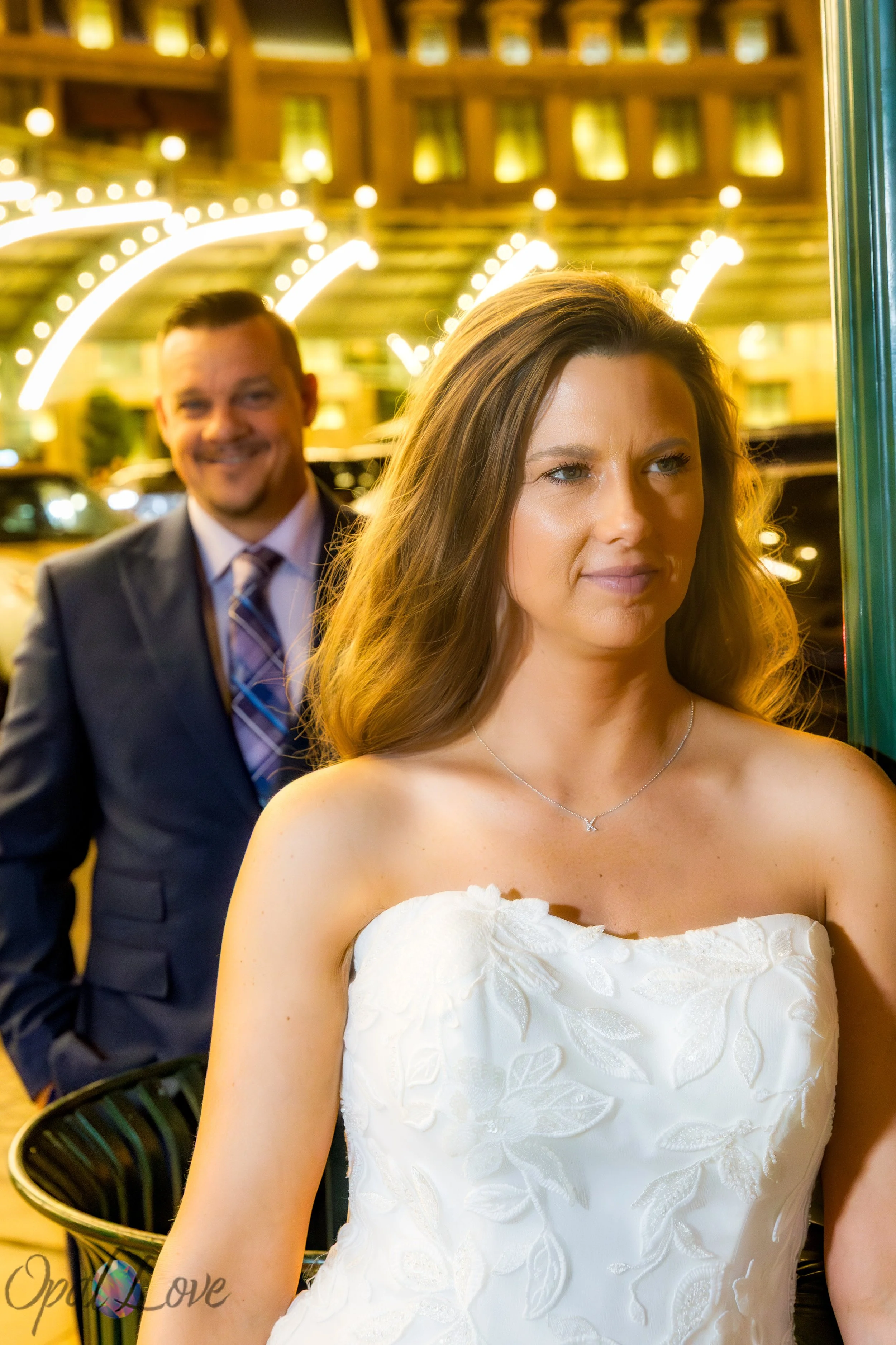 Bride in focus with groom smiling behind her under warm string lights at Paris Las Vegas.