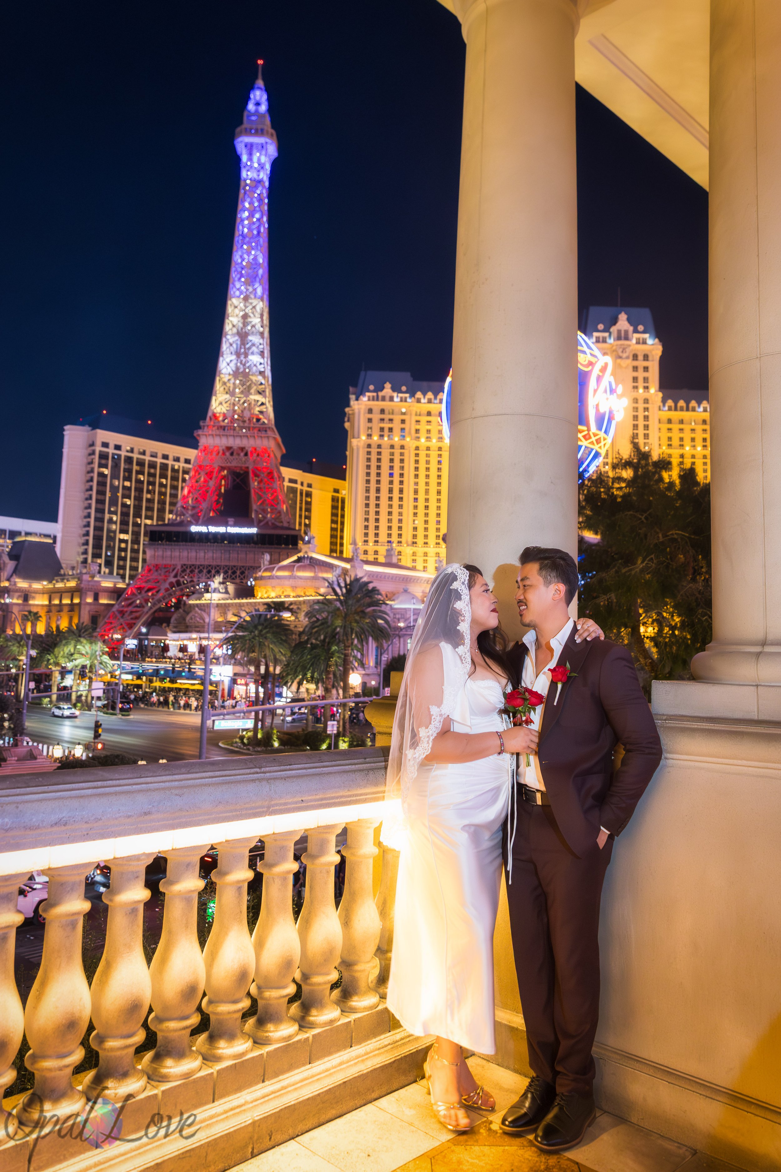 Couple embracing on the Bellagio balcony overlooking the Las Vegas Strip and the Paris Eiffel Tower at night.