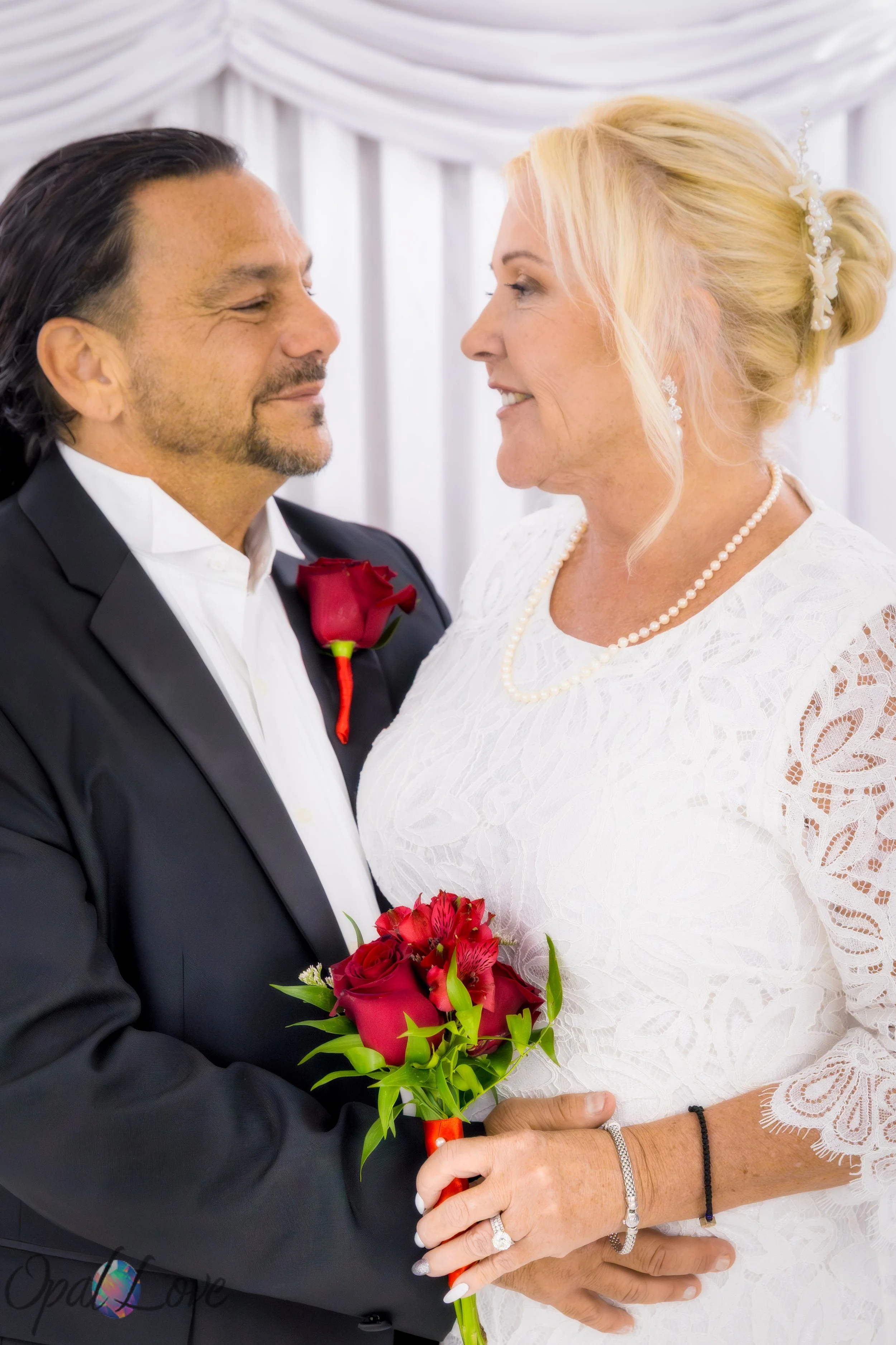 Newly married couple smiling at each other while holding a red rose bouquet inside the Little White Wedding Chapel.