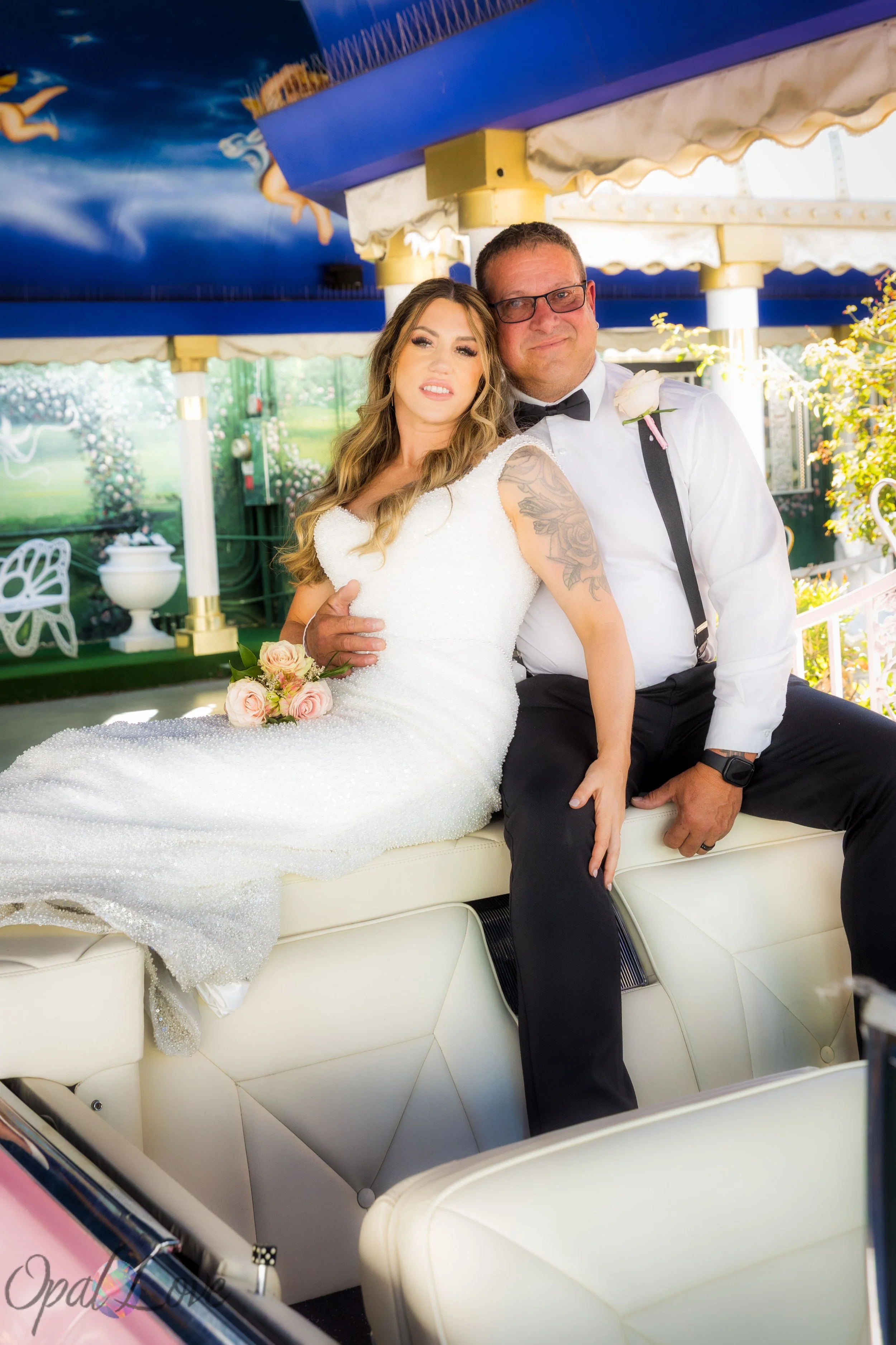 Newlyweds seated together in a pink Cadillac beneath a blue painted ceiling.