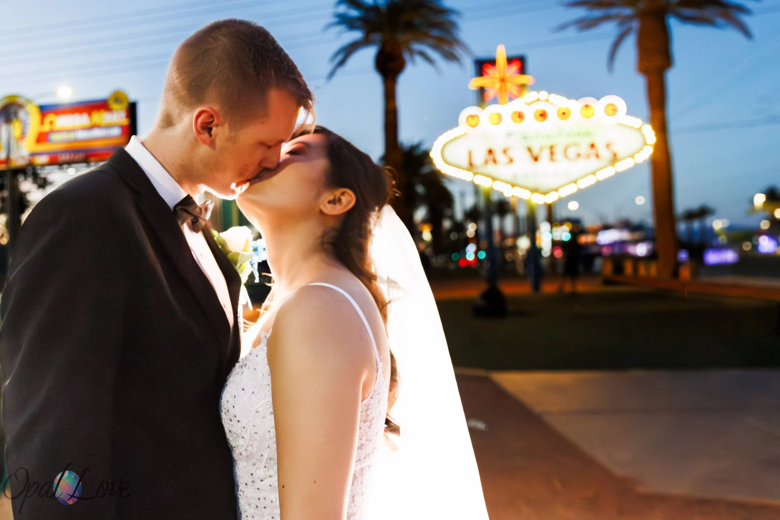 Newlywed couple kissing in front of the glowing Las Vegas sign at night.