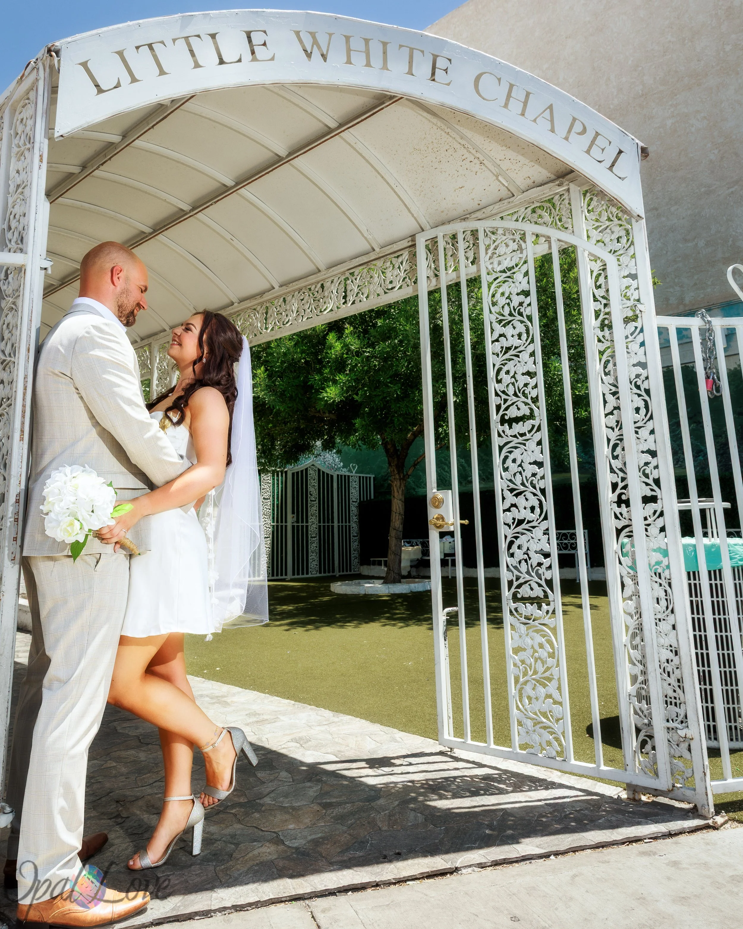 Couple embracing beneath the Little White Chapel garden arch, with the bride lifting one heel.