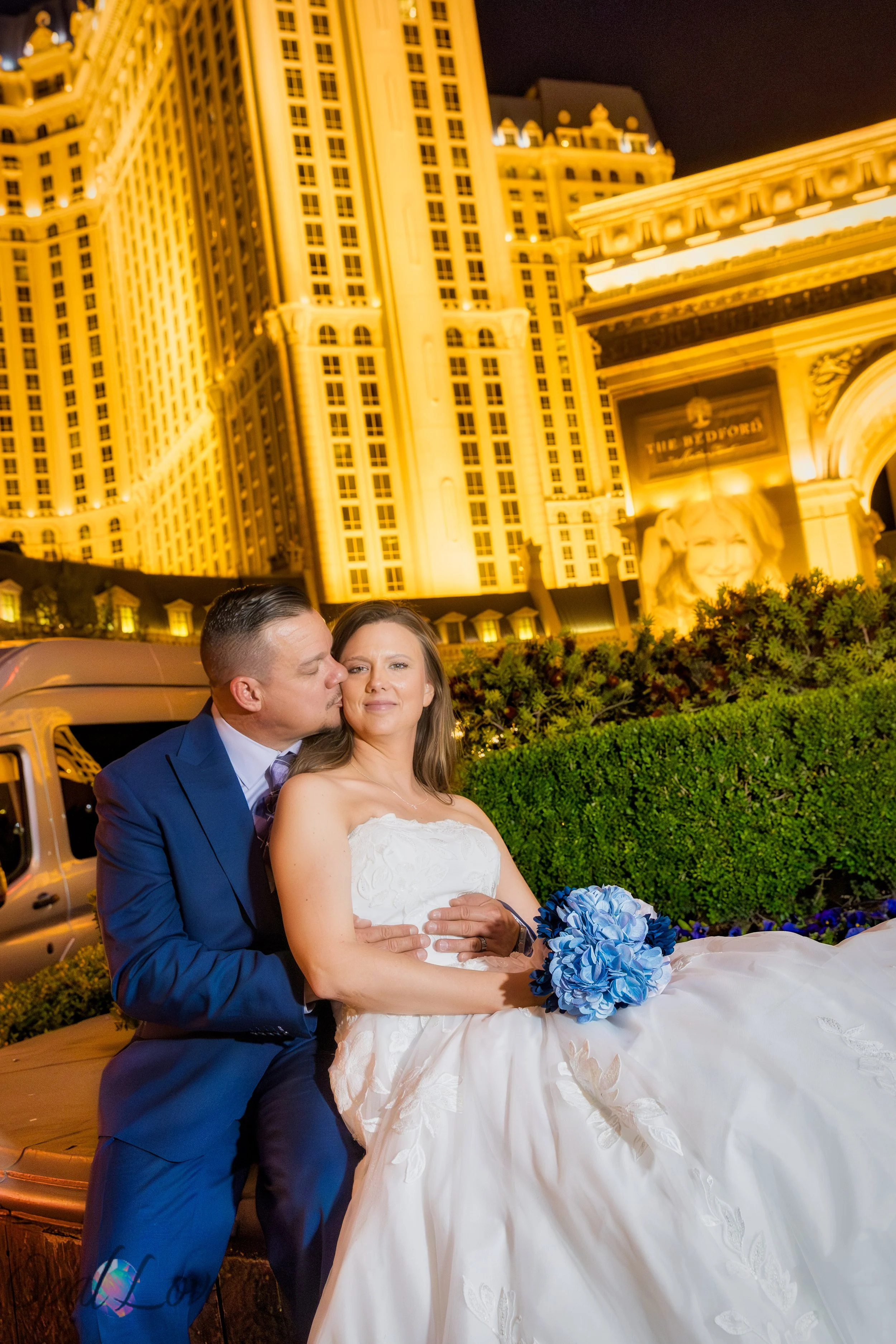 Bride and groom sitting together in front of Paris Las Vegas glowing buildings at night.