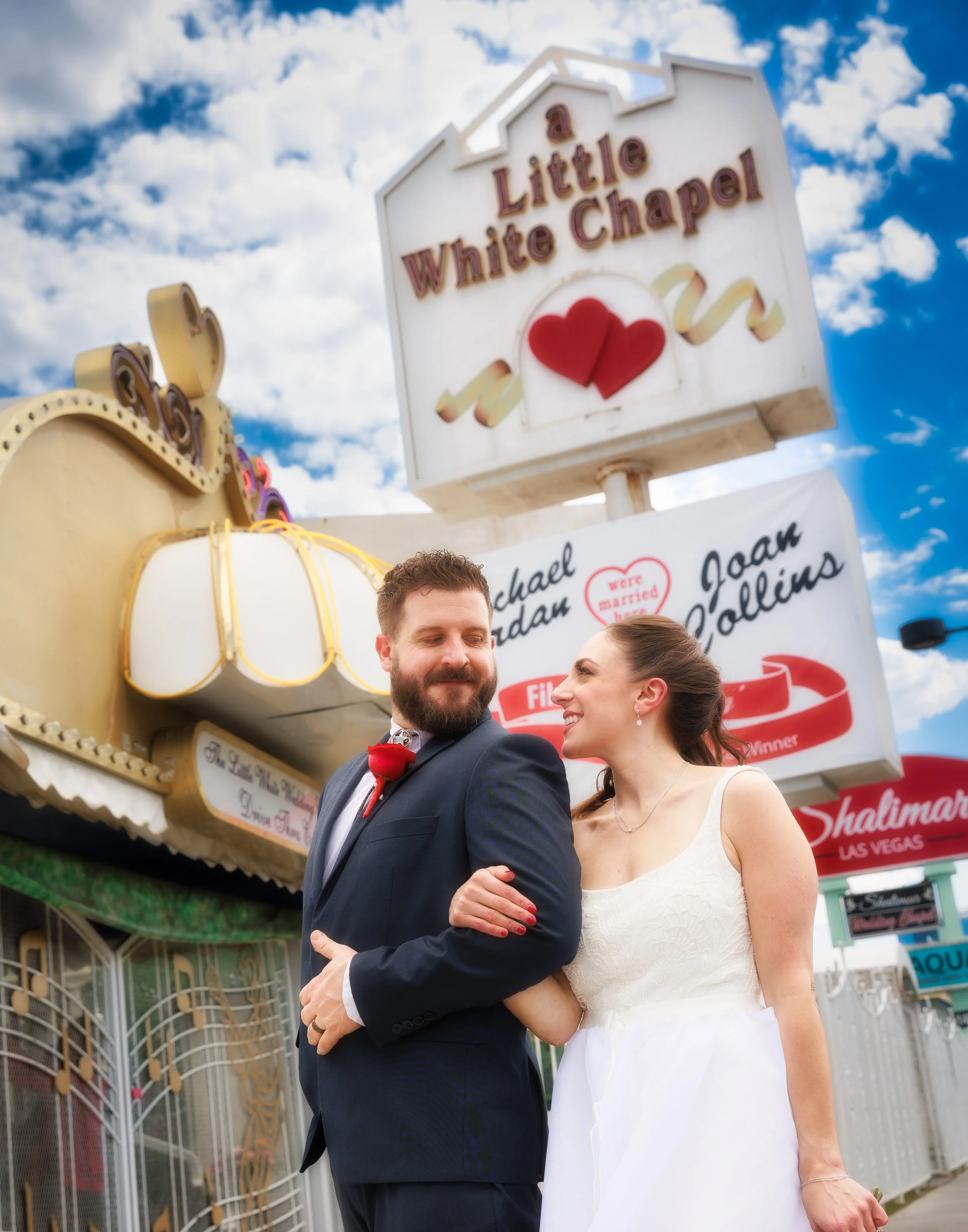 Newlyweds walking together beneath the Little White Chapel sign on a sunny Las Vegas day.