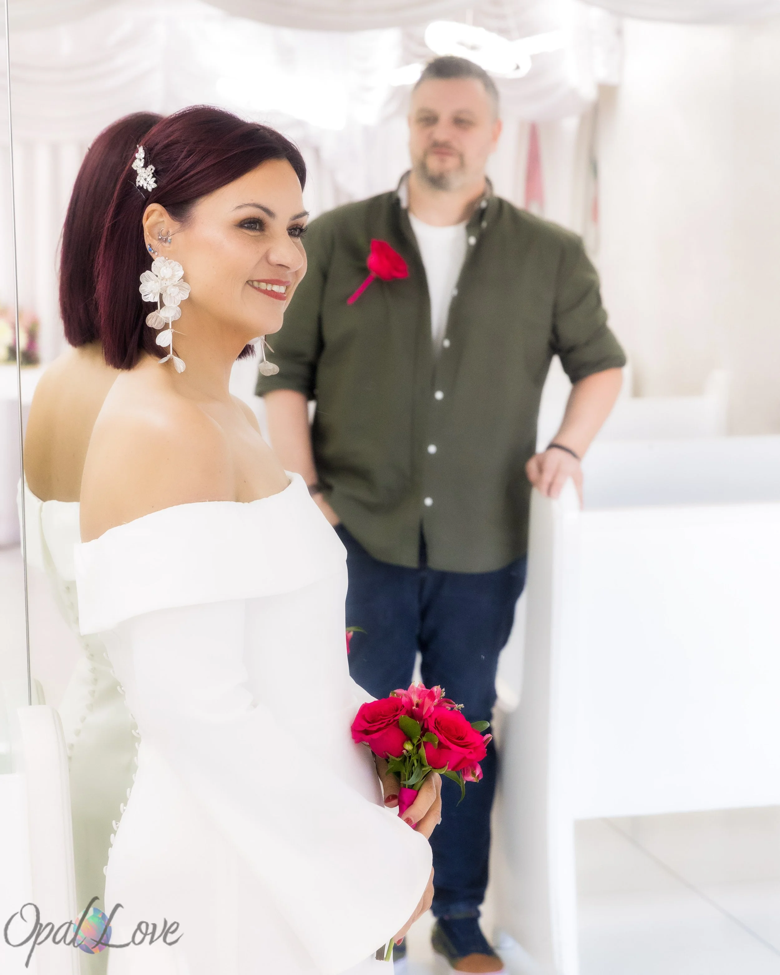 Bride holding a red rose bouquet with groom standing behind her inside the chapel.