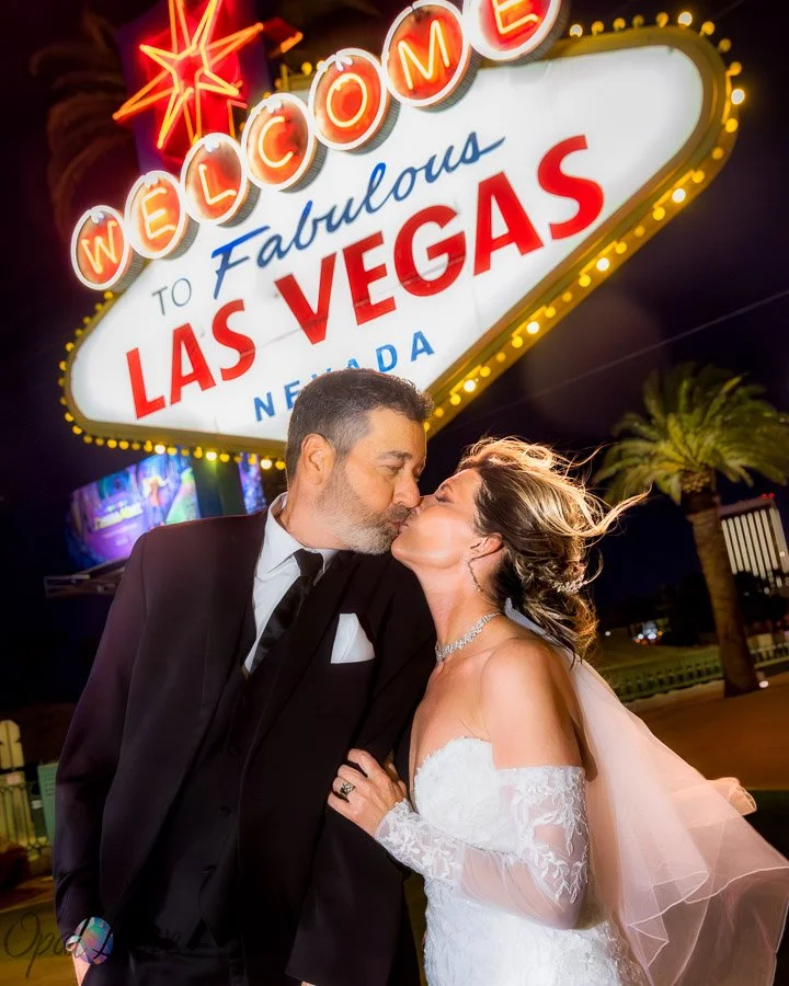 Newlywed couple kissing beneath the Welcome to Fabulous Las Vegas sign at night.
