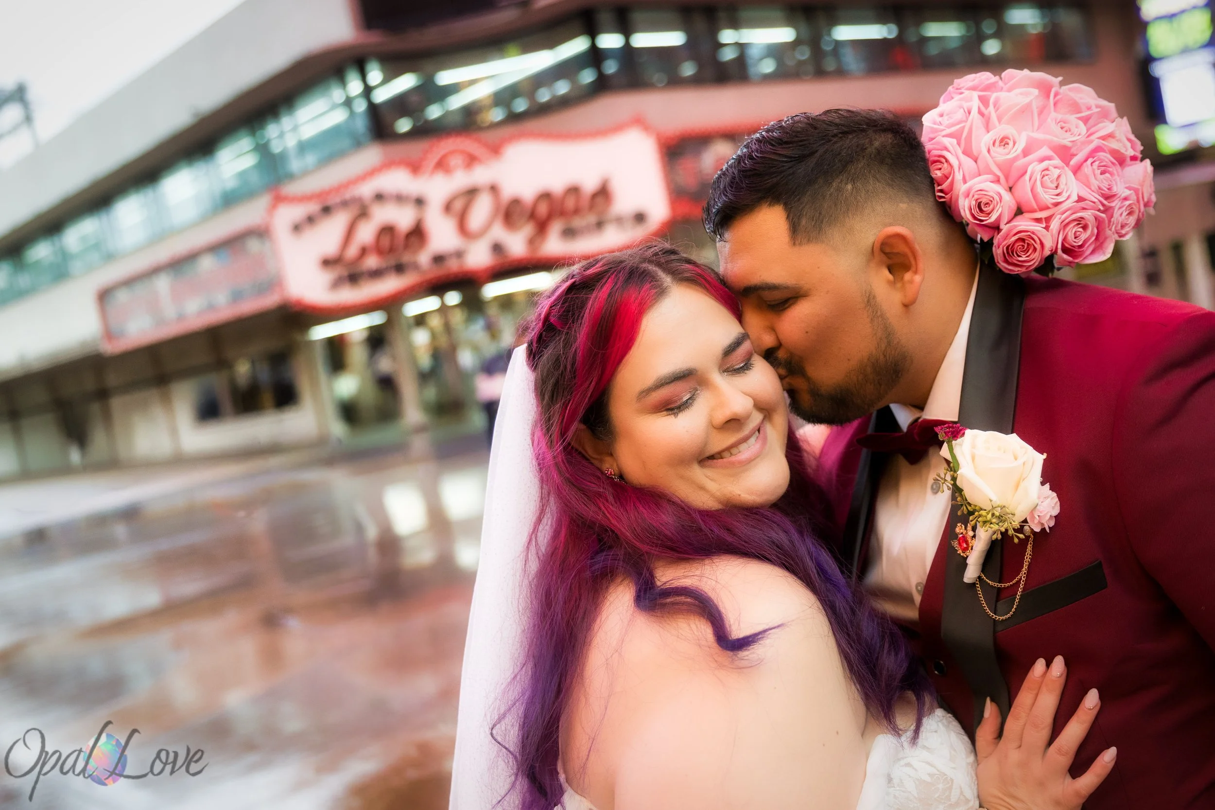 Groom kissing bride’s cheek with vintage Las Vegas signage blurred behind them.