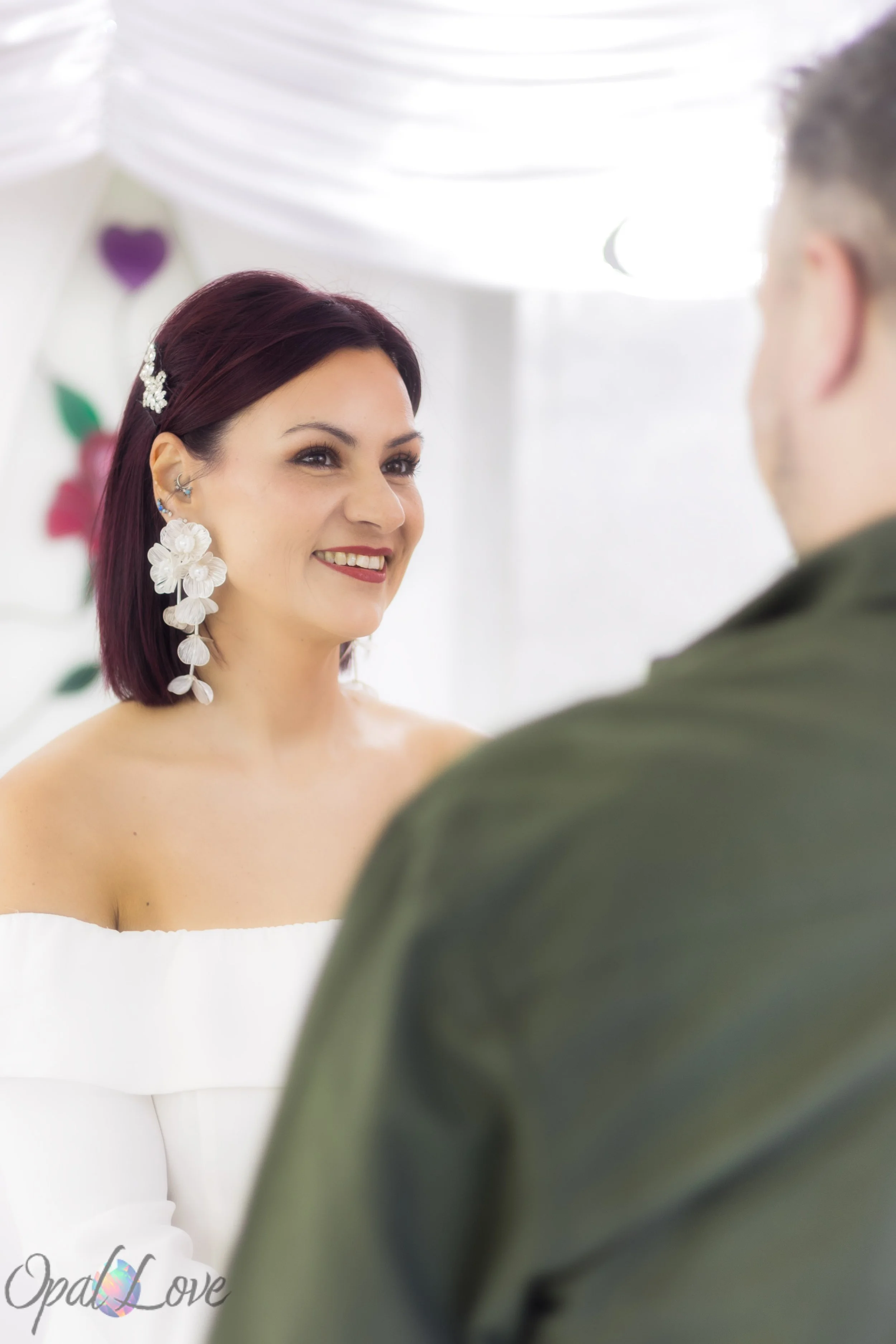 Bride smiling at her partner during their ceremony, wearing large white floral earrings.