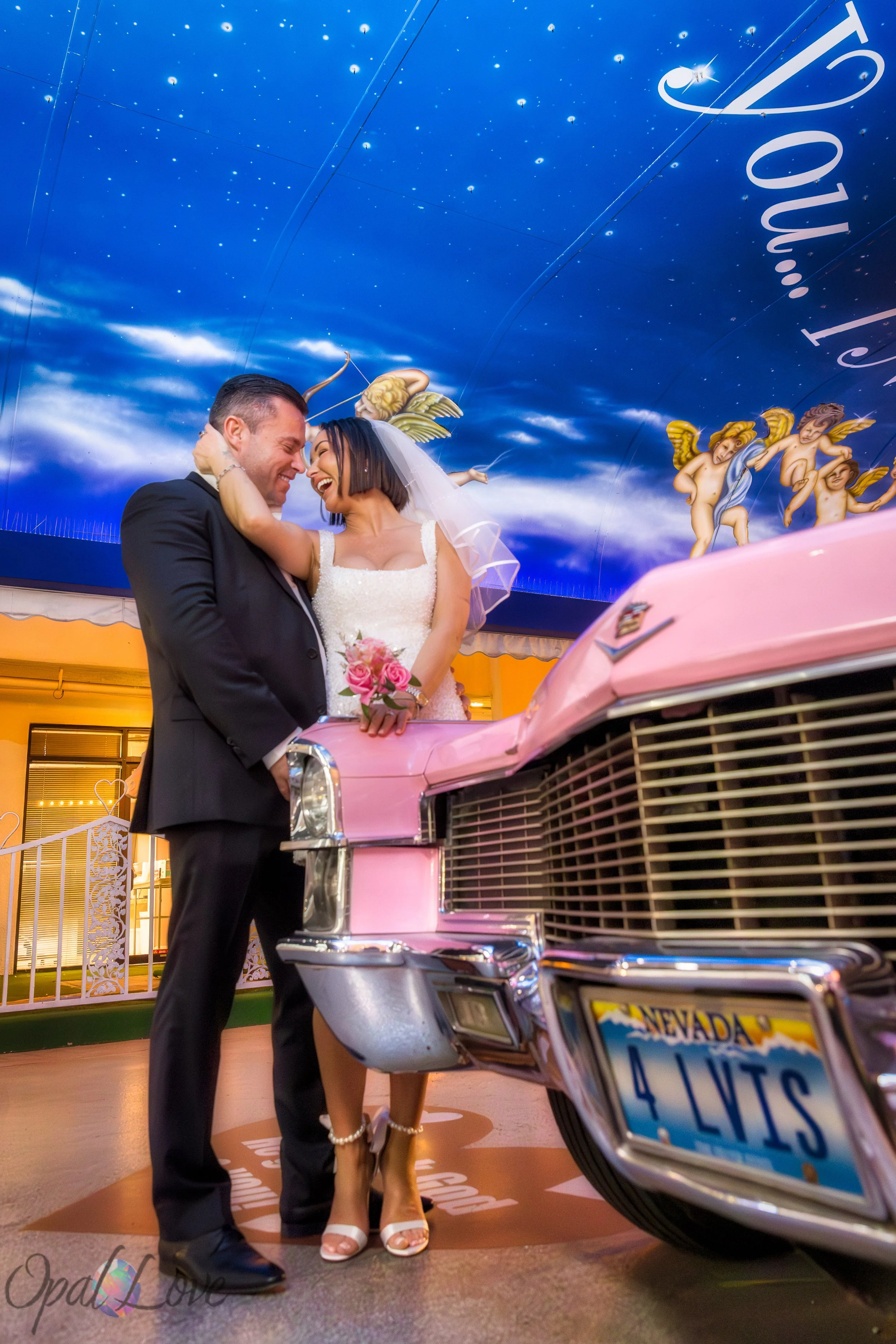 Bride and groom laughing beside the pink Cadillac at Little White Wedding Chapel at night.