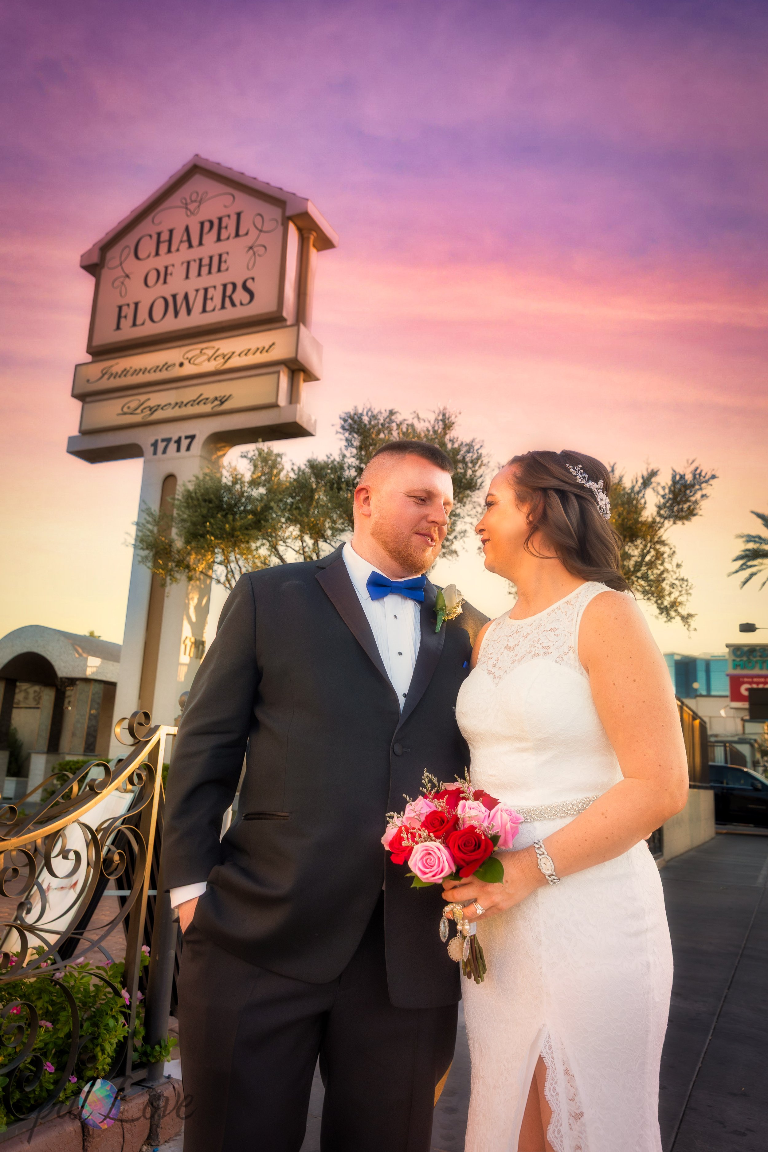 Newlyweds smiling at each other beneath the Chapel of the Flowers sign at sunset.