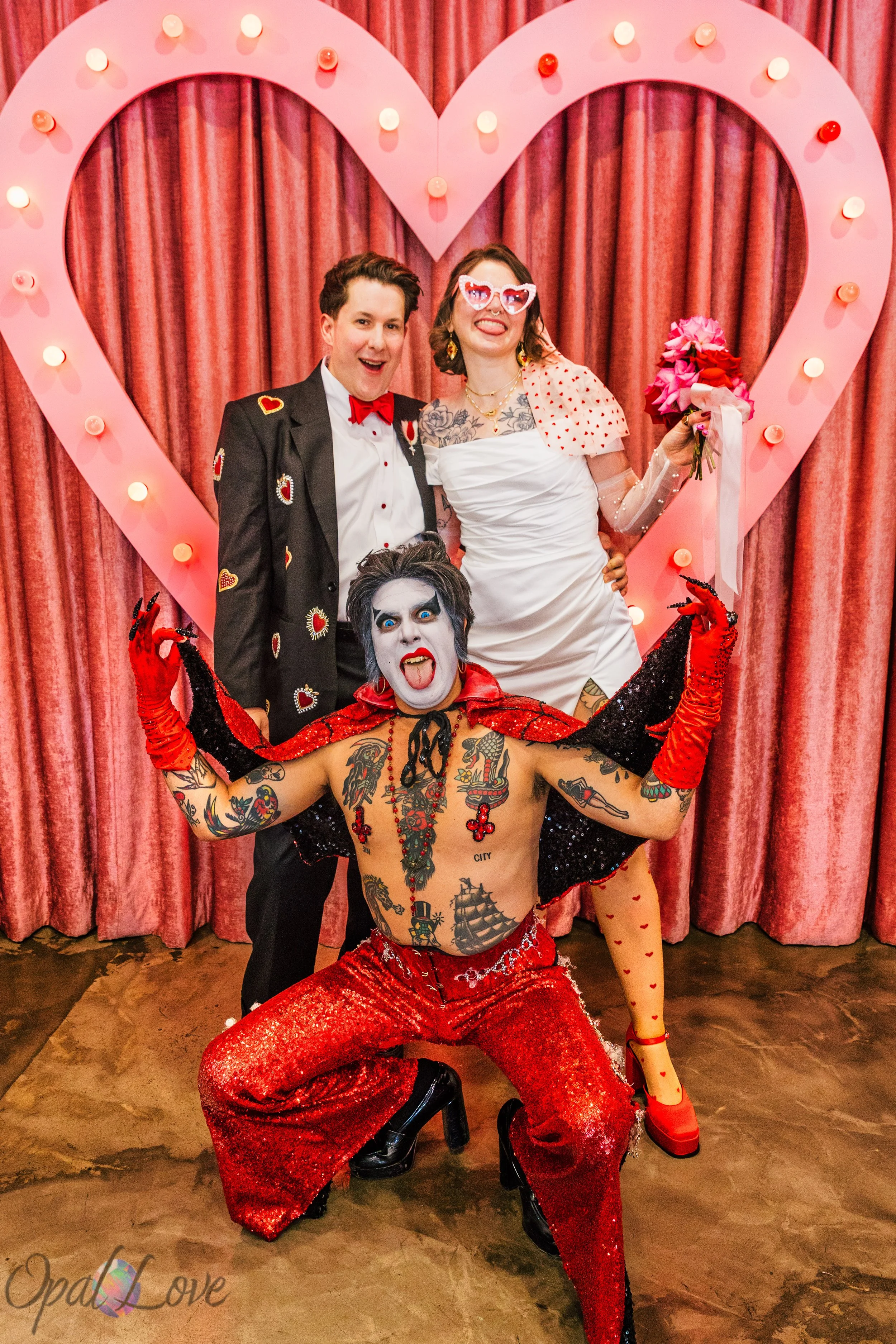 Newlyweds Rachel and Tom pose with Hellvis beneath the illuminated heart backdrop at A Sure Thing Wedding Chapel.