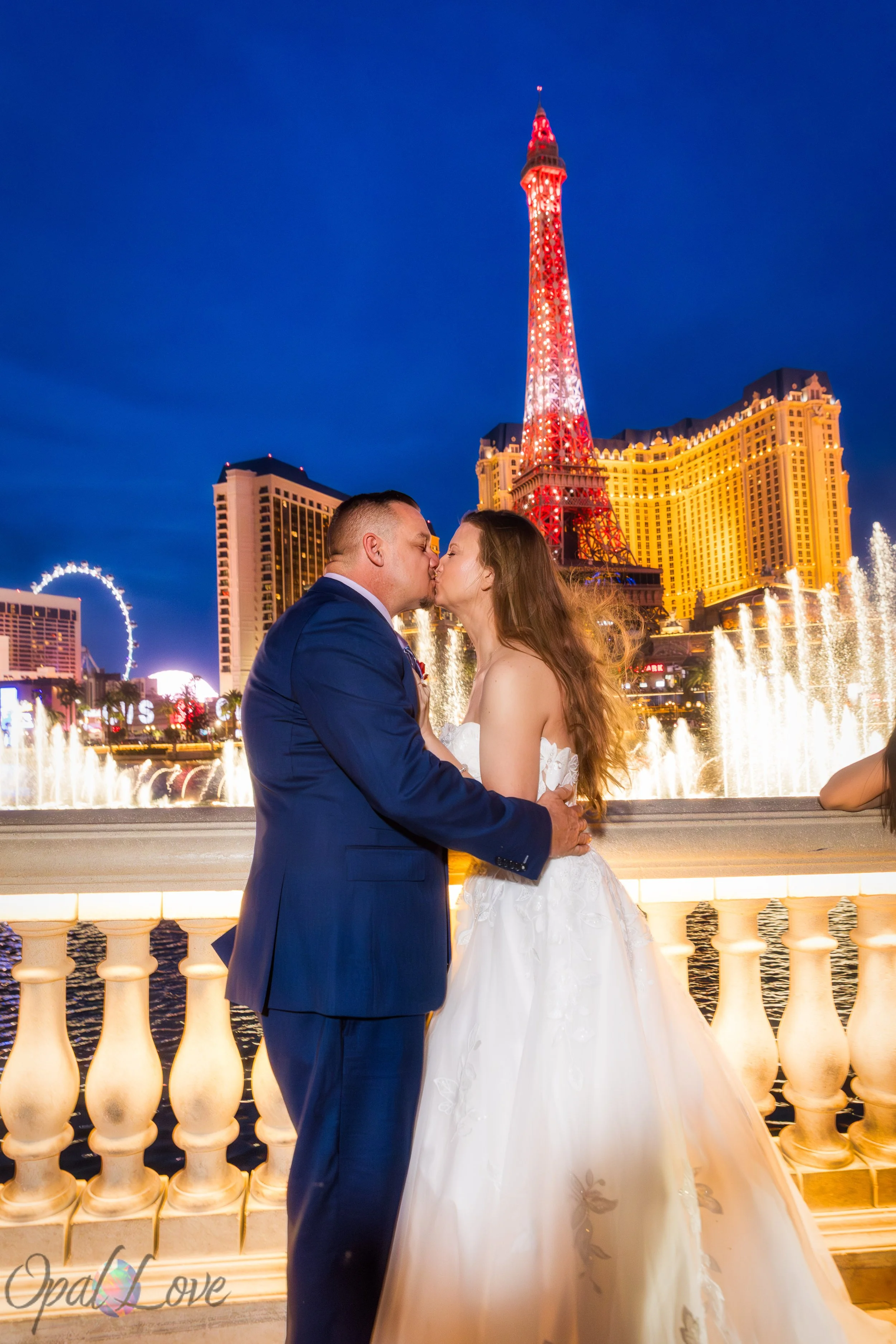 Couple sharing a kiss with the Eiffel Tower from Paris Las Vegas visible behind them across the Bellagio fountains.