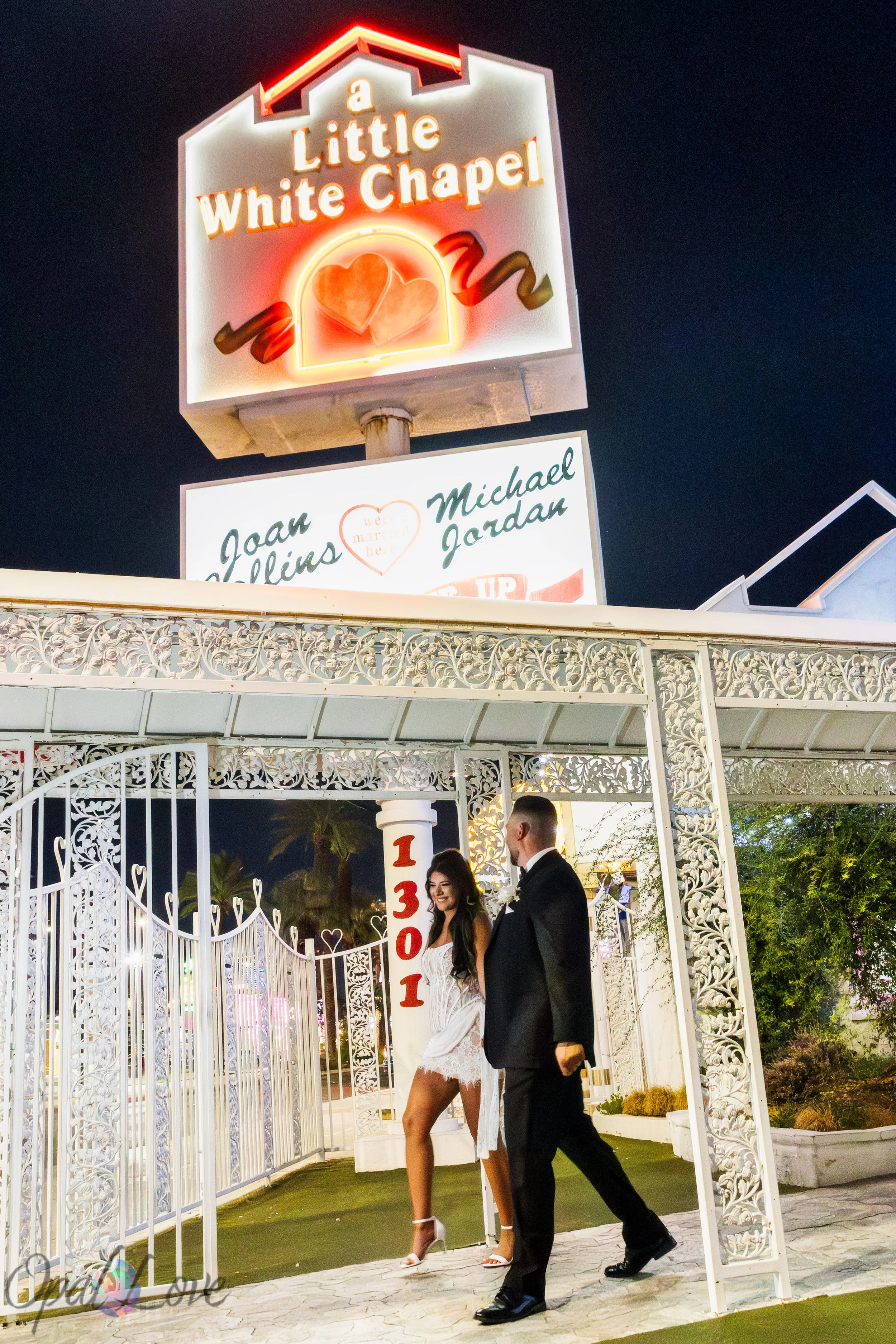 Alysha and Justin walking beneath the white ironwork and neon sign at the Little White Wedding Chapel in Las Vegas.