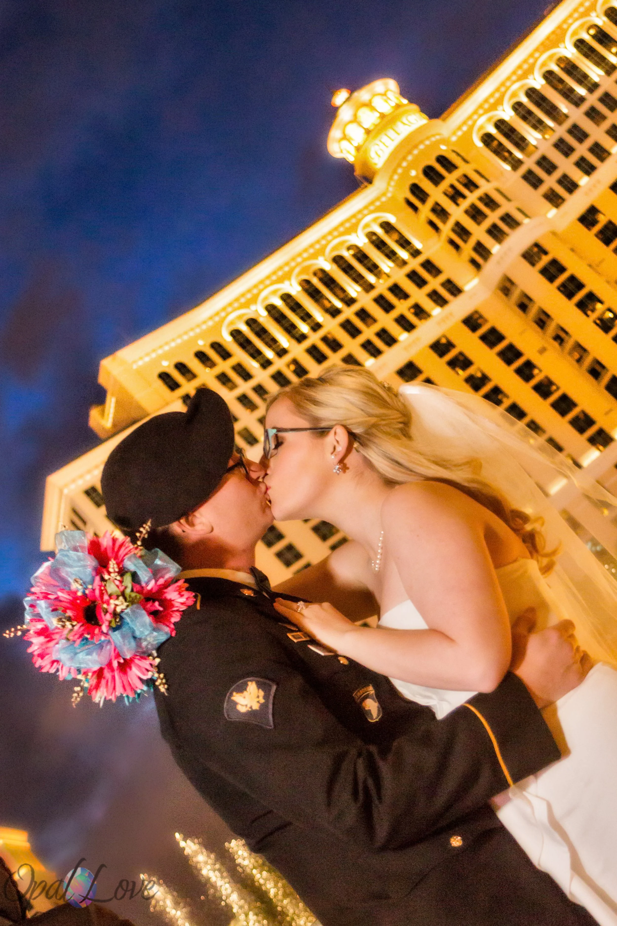 Bride kissing a groom in military uniform beneath glowing Las Vegas hotel lights.