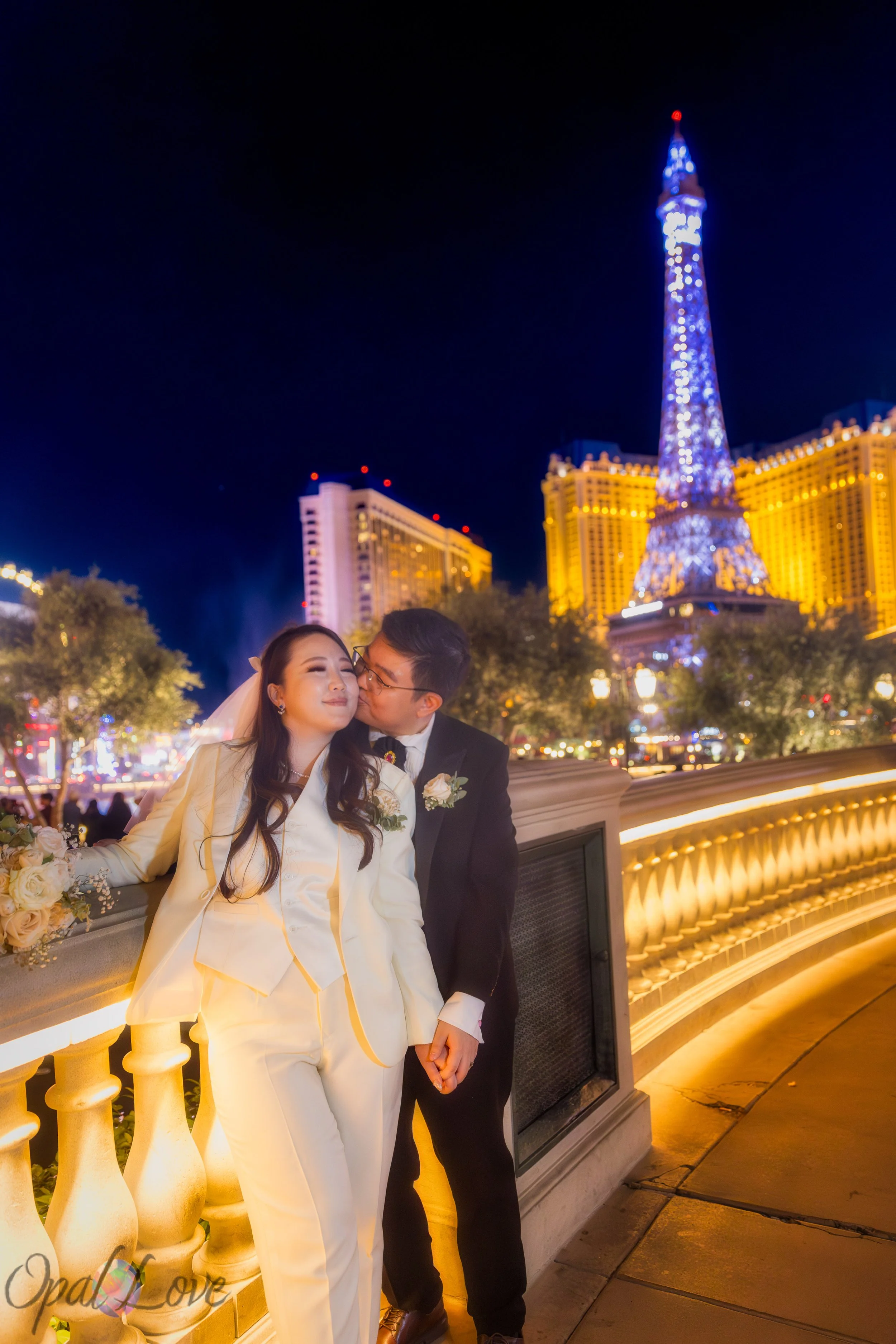 Bride and groom posing on bridge with Eiffel Tower lit up at night in Las Vegas