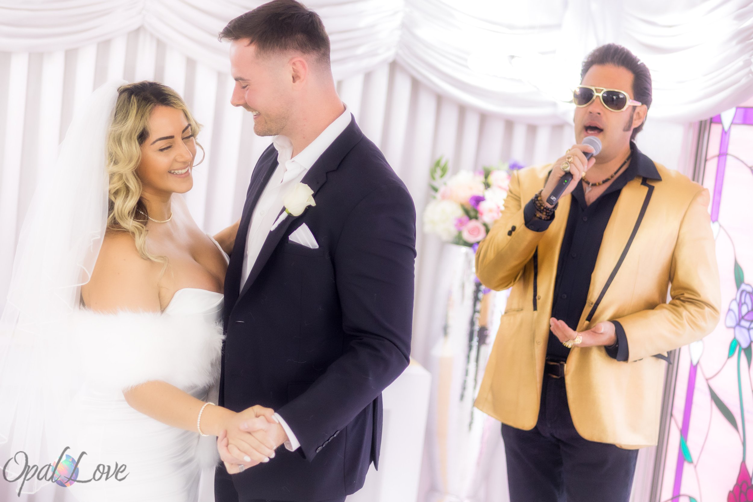 Couple holding hands in a white chapel while an Elvis impersonator sings beside them.