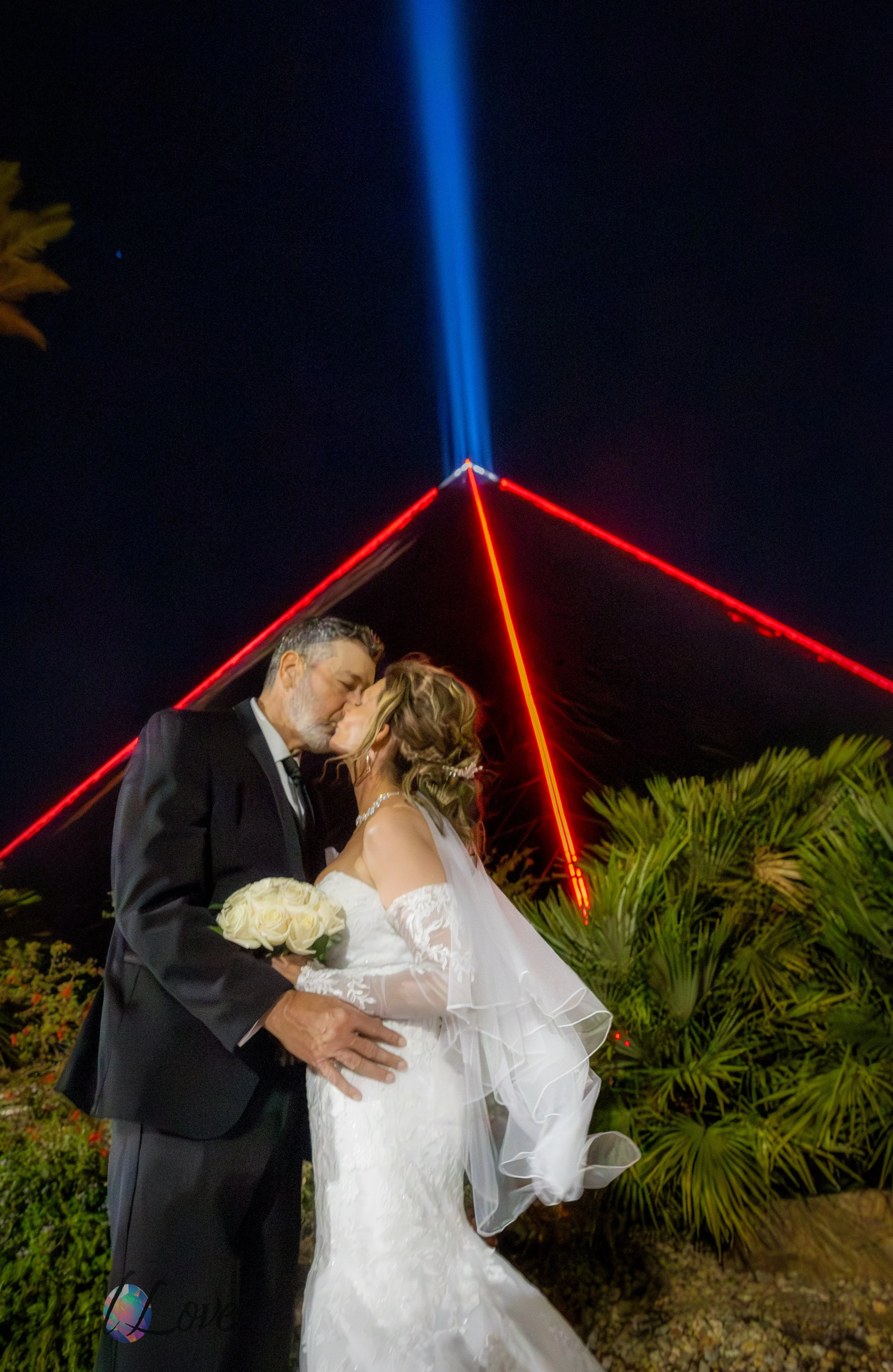 Couple kissing under the Luxor pyramid beam at night surrounded by palm trees.