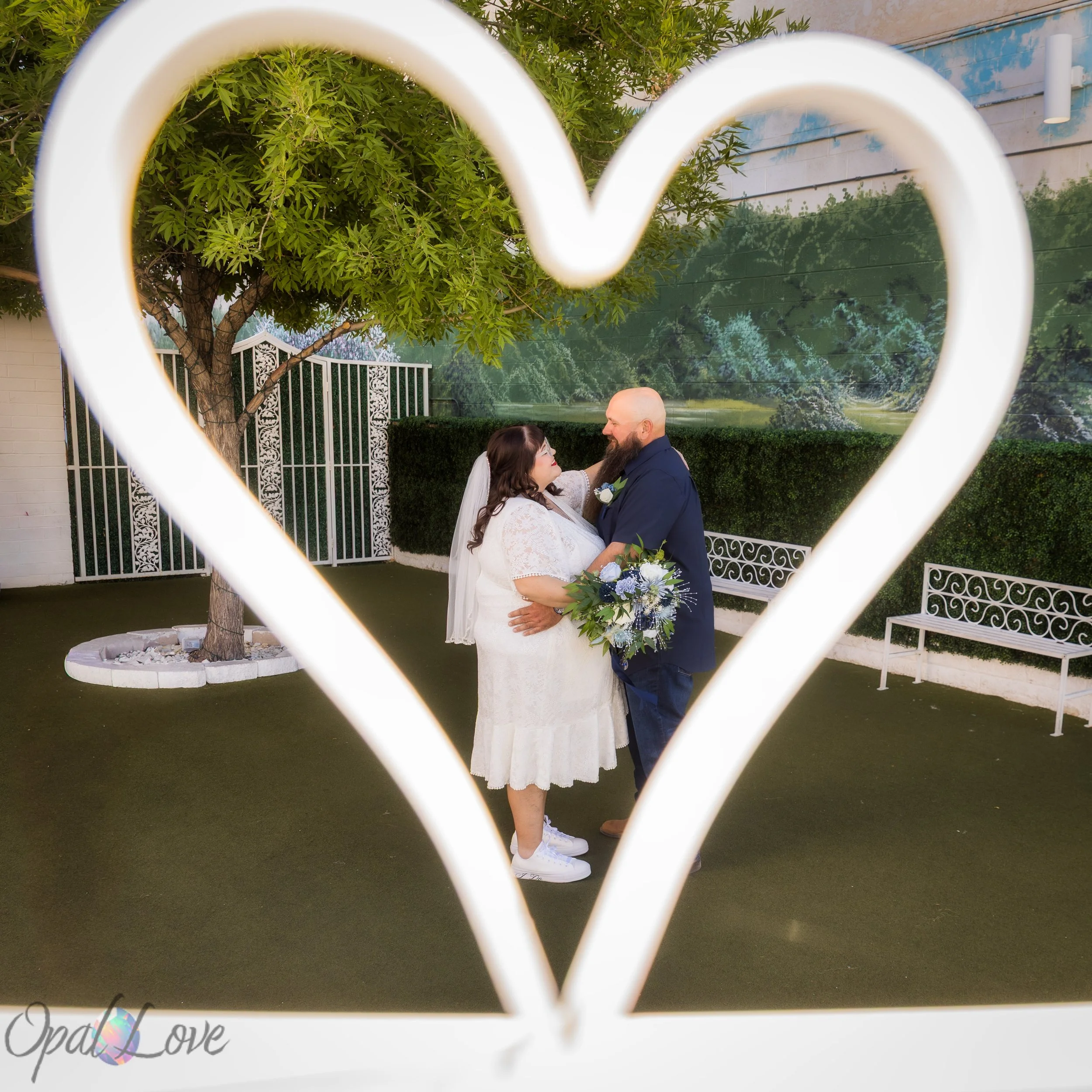 Couple holding each other while framed through a white heart sculpture in the chapel courtyard