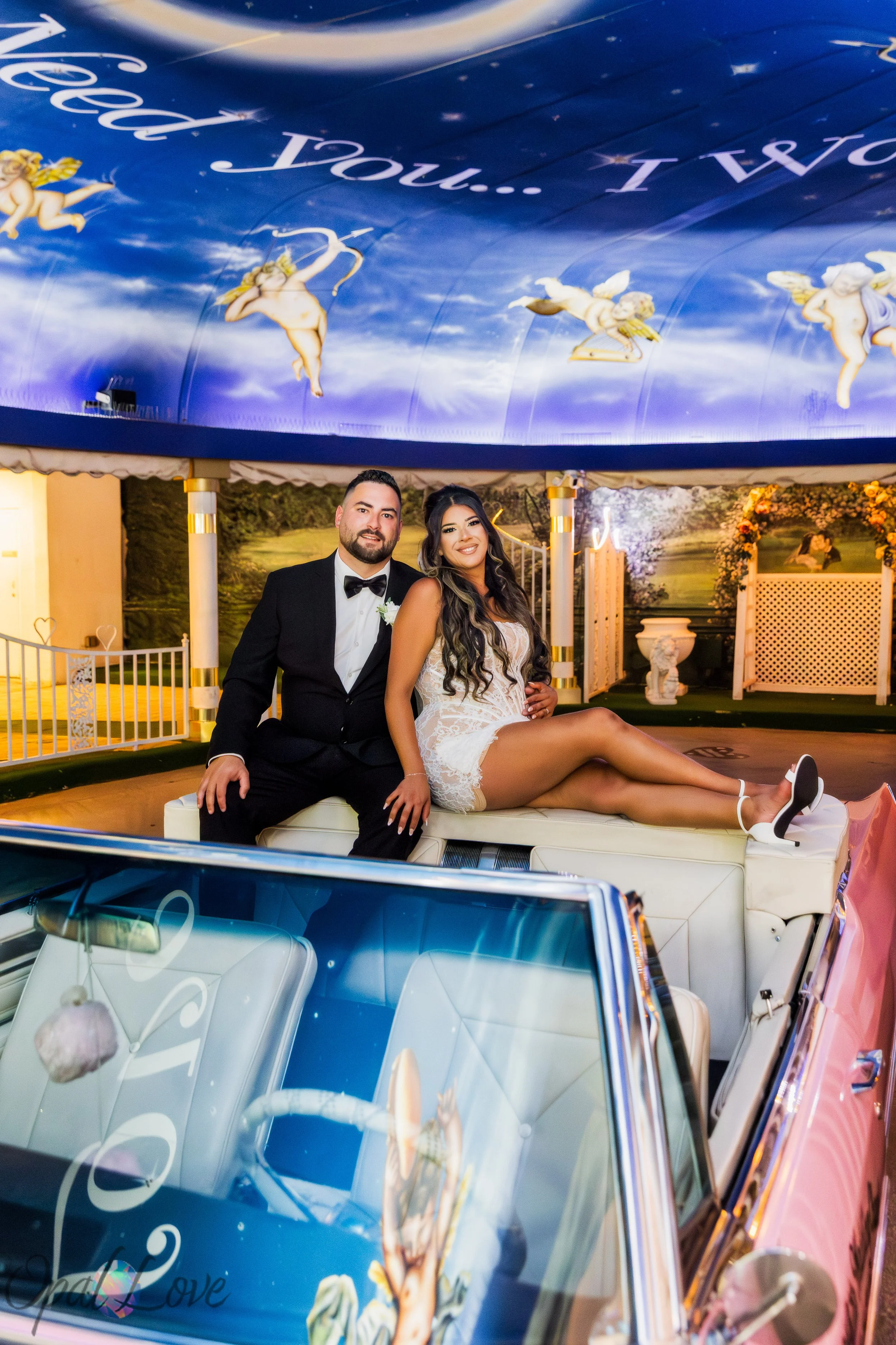 Alysha and Justin posing together on the pink Cadillac beneath the blue cherub ceiling at the Little White Wedding Chapel.