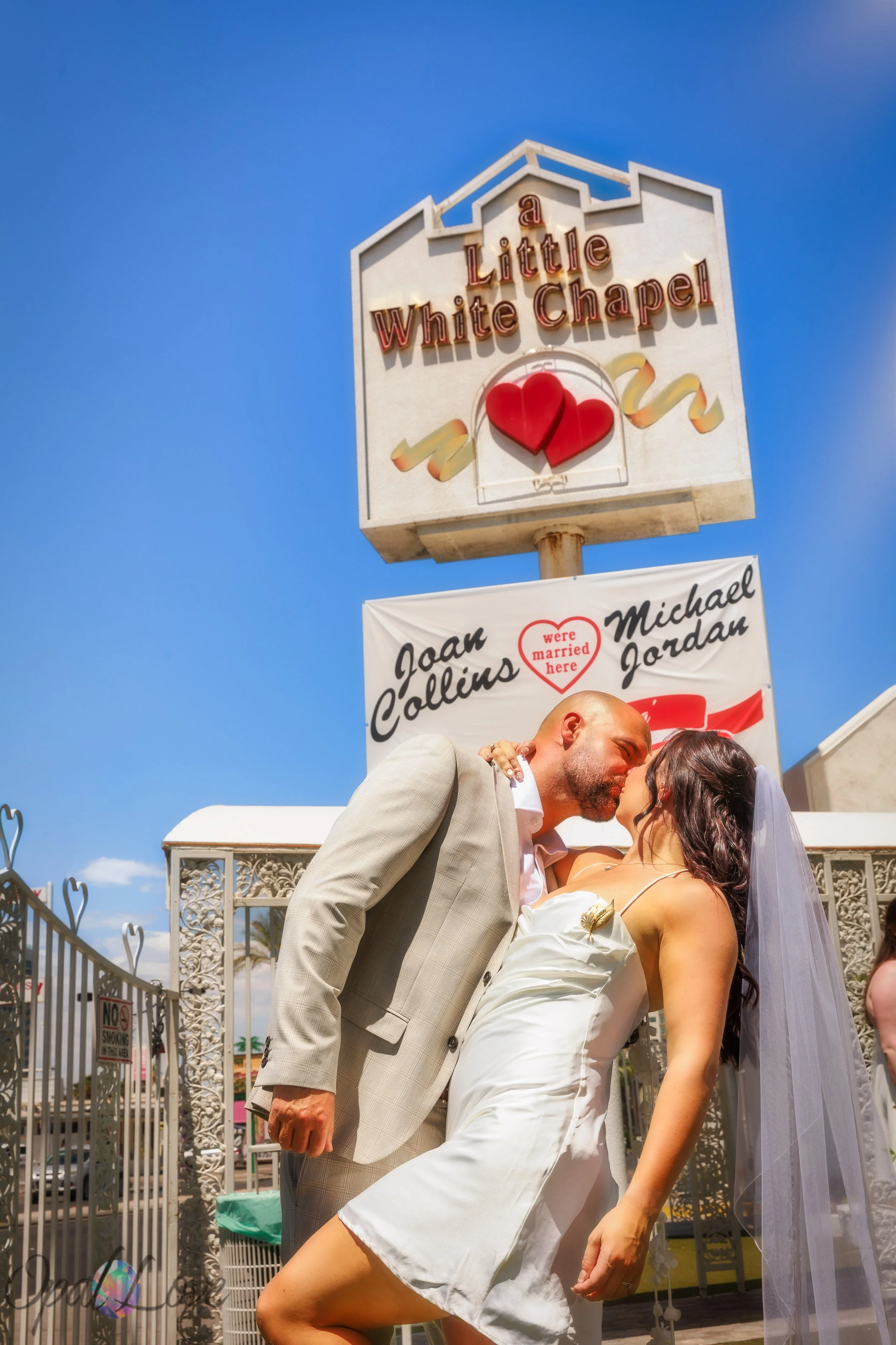 Newlyweds kissing beneath the Little White Wedding Chapel sign against a bright blue sky.