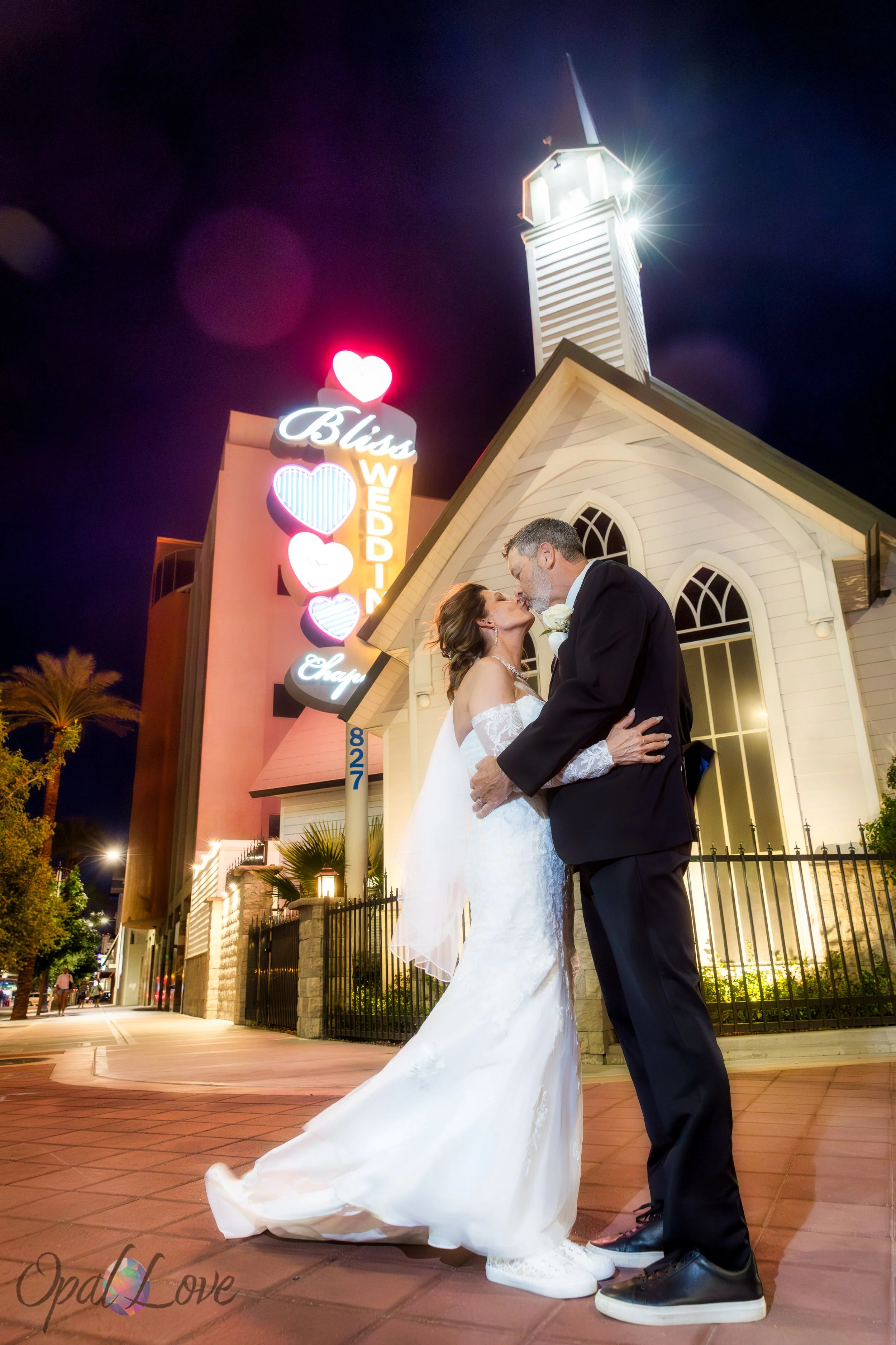 Couple kissing in front of Bliss Wedding Chapel sign at night, neon hearts glowing behind them.